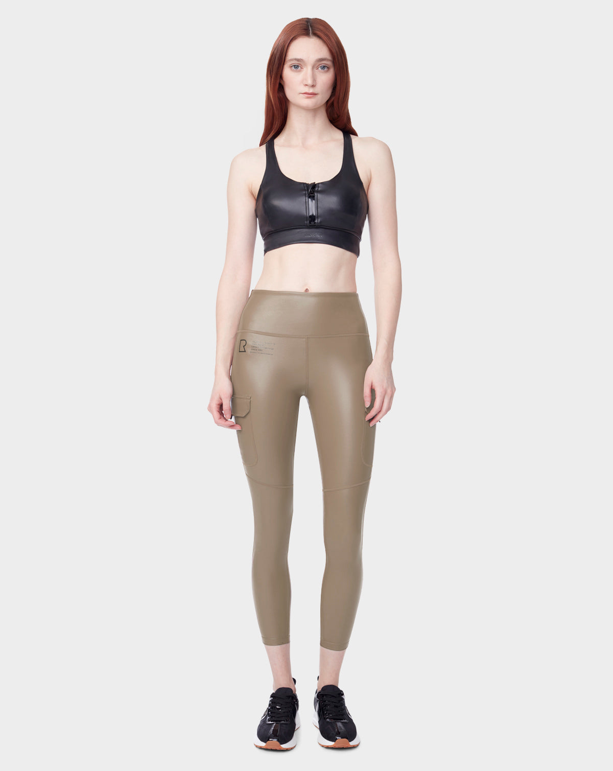 A woman faces forward in Rudsak’s SIEN L - 8523010 Cement: a black zip-front sports bra and high-waisted tan cargo leggings with side pocket, paired with black sneakers, all set against a light background.
