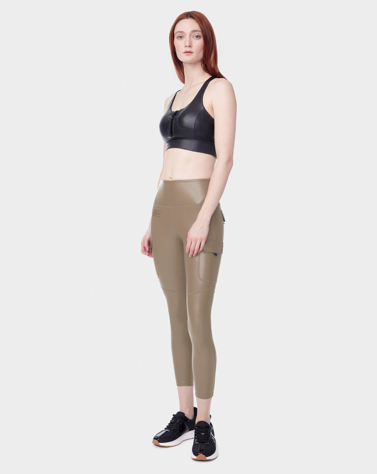 A woman with long red hair stands against a plain background, wearing the Rudsak SIEN L - 8523010 Cement cargo leggings from womens activewear, a black sports bra, and black sneakers. She faces forward with a neutral expression.
