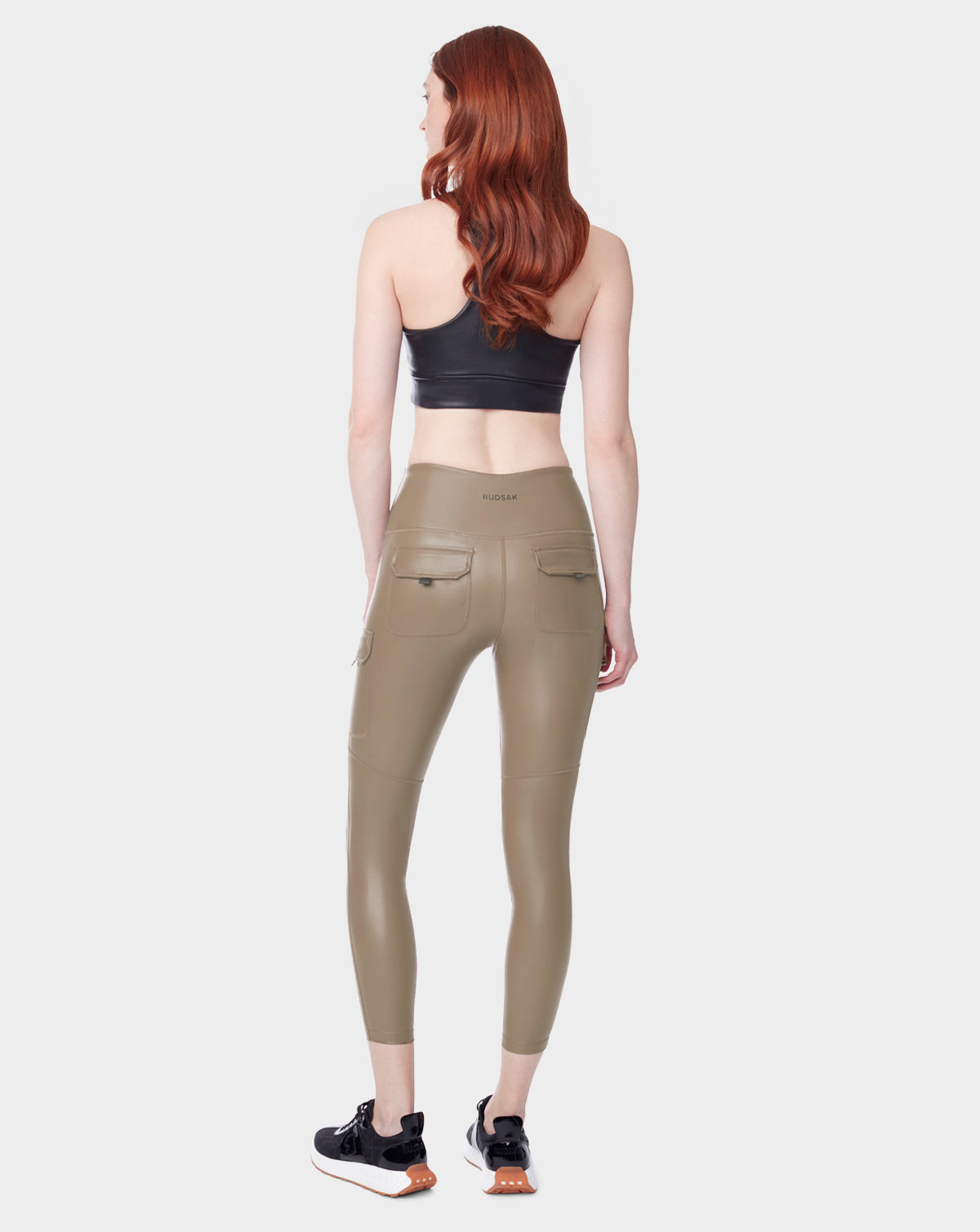 A woman with long red hair, wearing a black crop top and olive green cargo leggings with back pockets, stands facing away from the camera. She completes her look with Rudsaks SIEN L - 8523010 Cement sneakers in black and white.