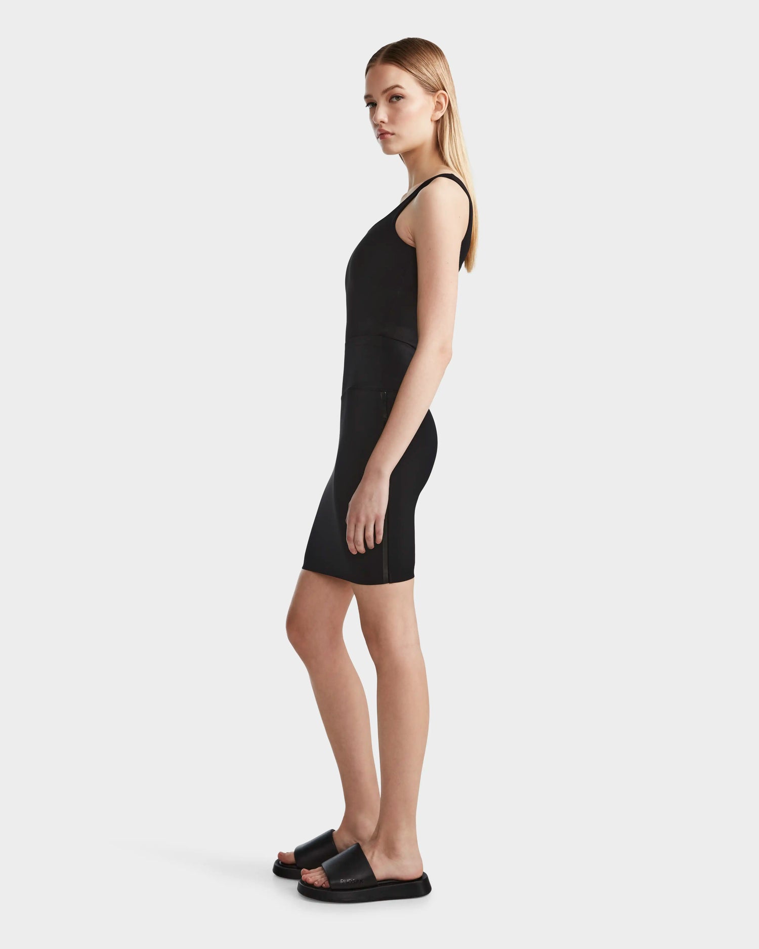 A woman with straight blonde hair wears the JESS - 8523042 BLACK fitted tank top and bike shorts by Rudsak, paired with black slide sandals, creating an effortless spring look against a plain white background.
