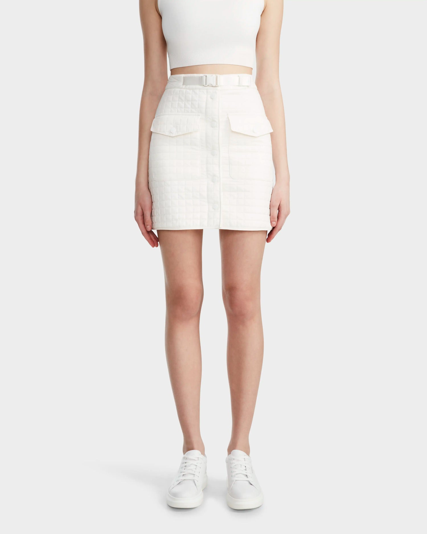 A woman wears the Rudsak SABINE - 8524063 Eggshell sleeveless crop top, a quilted short skirt with large pockets and a matching belt with buckle webbing front closure, paired with white sneakers against a light background.