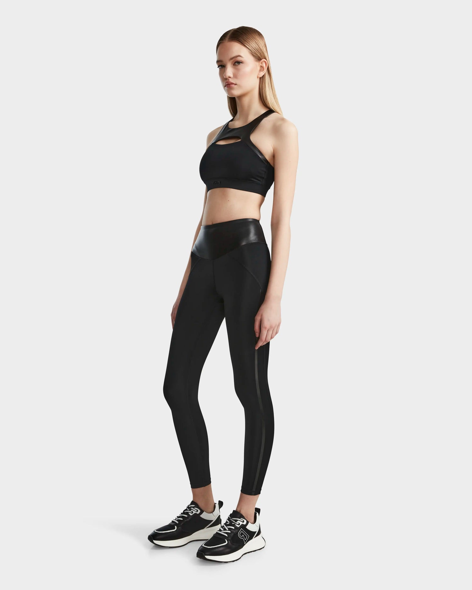 A woman with straight light brown hair and a neutral expression wears the RUDSAK ARYA - 8523002 BLACK sports bra and high-waisted leggings with glossy vegan leather panels, crafted from MVMT+ fabric, paired with black sneakers featuring white soles.