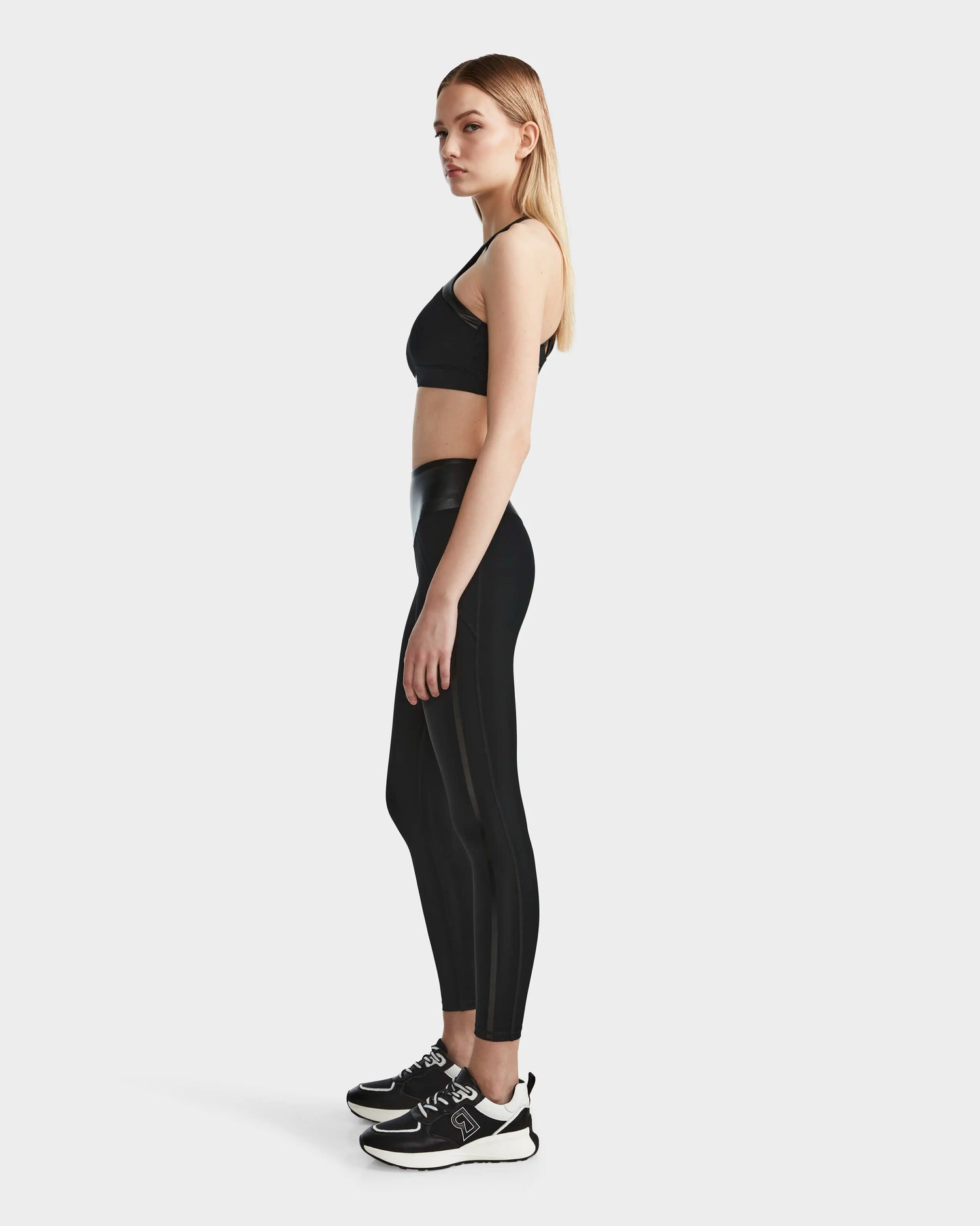 A woman with straight blonde hair in profile wears the RUDSAK ARYA - 8523002 BLACK sports bra and high-waisted leggings, paired with black sneakers with white soles, against a plain light background.