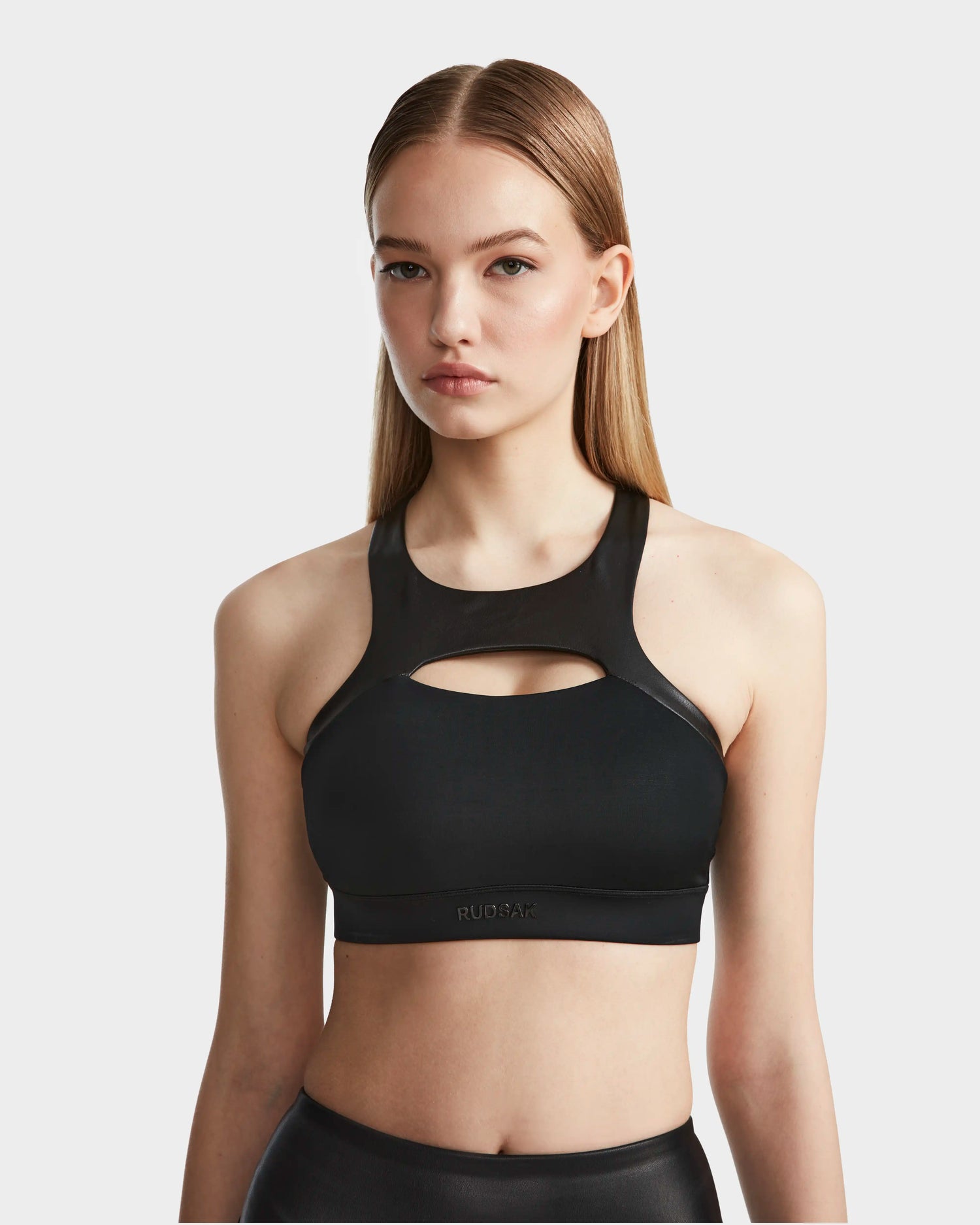 A woman with straight, light brown hair wears the RUDSAK ARYA - 8523002 BLACK sports bra, featuring a front cutout and matching black bottoms, standing against a plain light background.