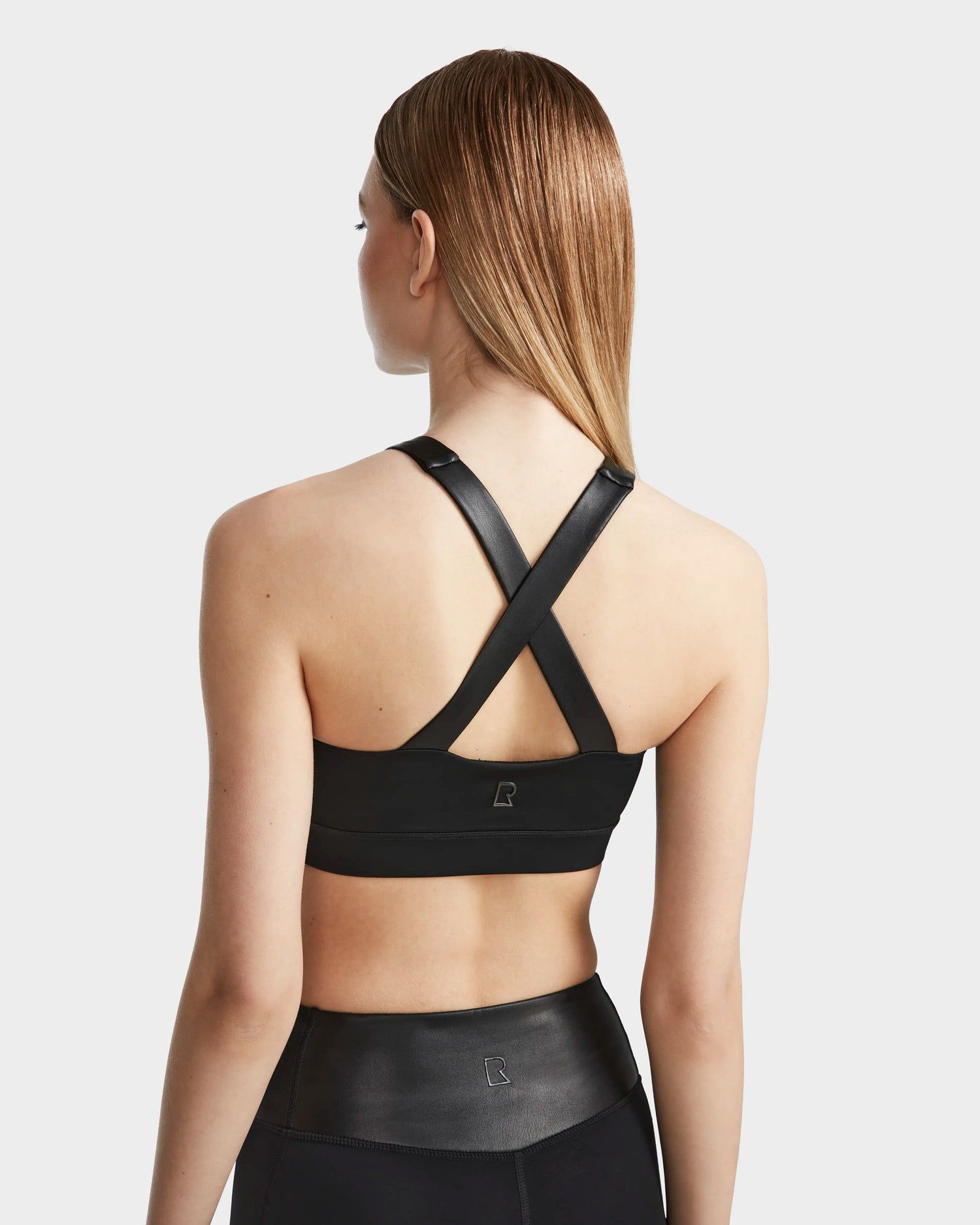 A woman with straight, light brown hair wears RUDSAKs ARYA - 8523002 BLACK sports bra featuring MVMT+ fabric and thick crisscrossed straps, paired with matching high-waisted leggings, standing against a plain white background.