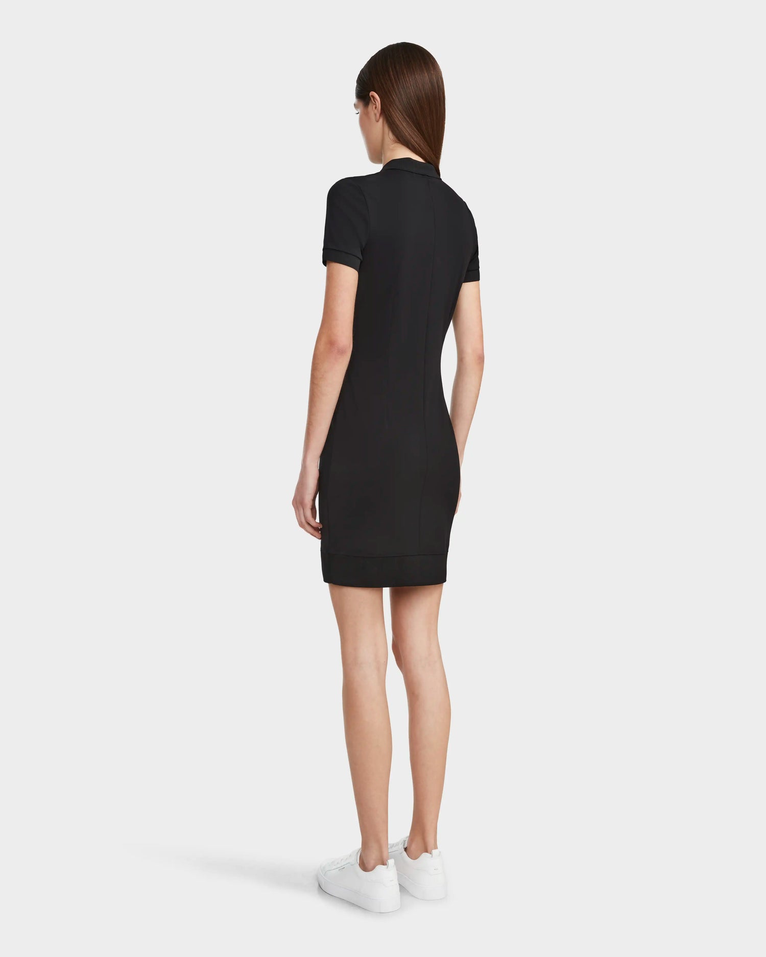A woman with straight brown hair stands facing away, highlighting sporty elegance in the Rudsak SUKI - 8524029 Black short-sleeve women's polo dress with white sneakers against a plain light background.