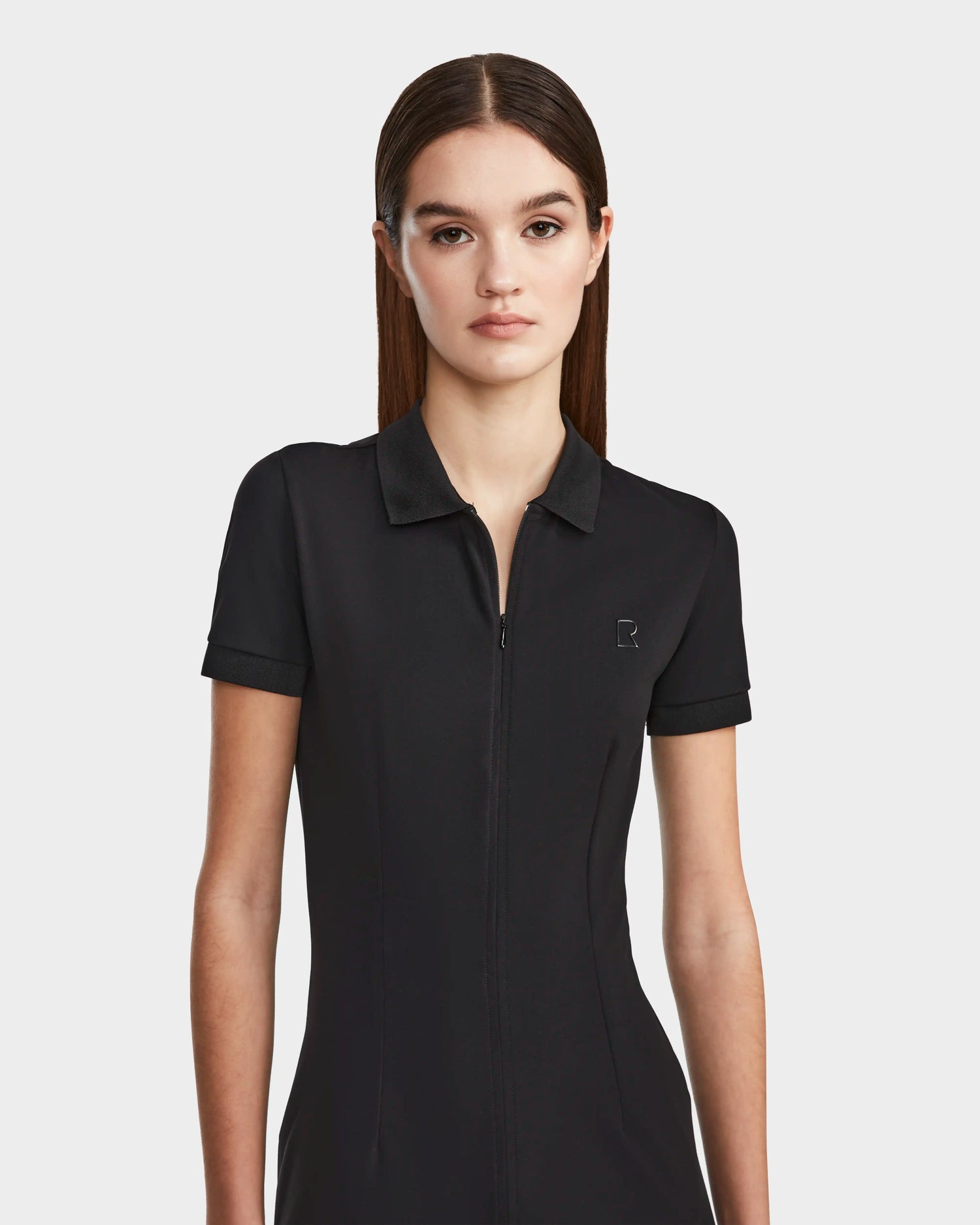 A woman with straight brown hair wears the Rudsak SUKI - 8524029 Black short-sleeve collared zip-up top, styled like a women’s polo dress, standing against a plain white background with a neutral expression.