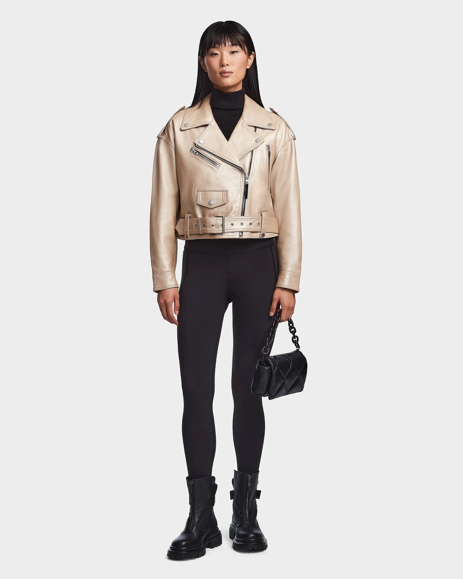 A woman faces forward, wearing the Rudsak MAGGIE - 8123031 Metallic Ivory women's leather biker jacket with a black turtleneck, black leggings, black combat boots, and holding a small black quilted handbag.