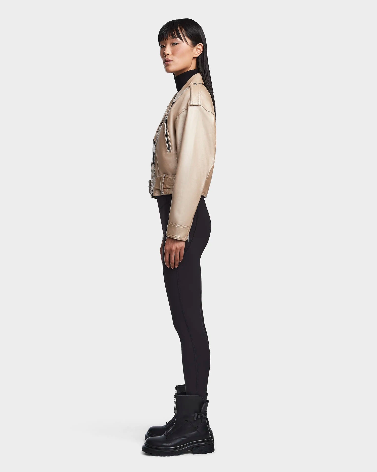 A woman with long straight hair stands in profile against a white background, wearing the Rudsak MAGGIE - 8123031 Metallic Ivory cropped leather biker jacket, black fitted pants, and black boots.