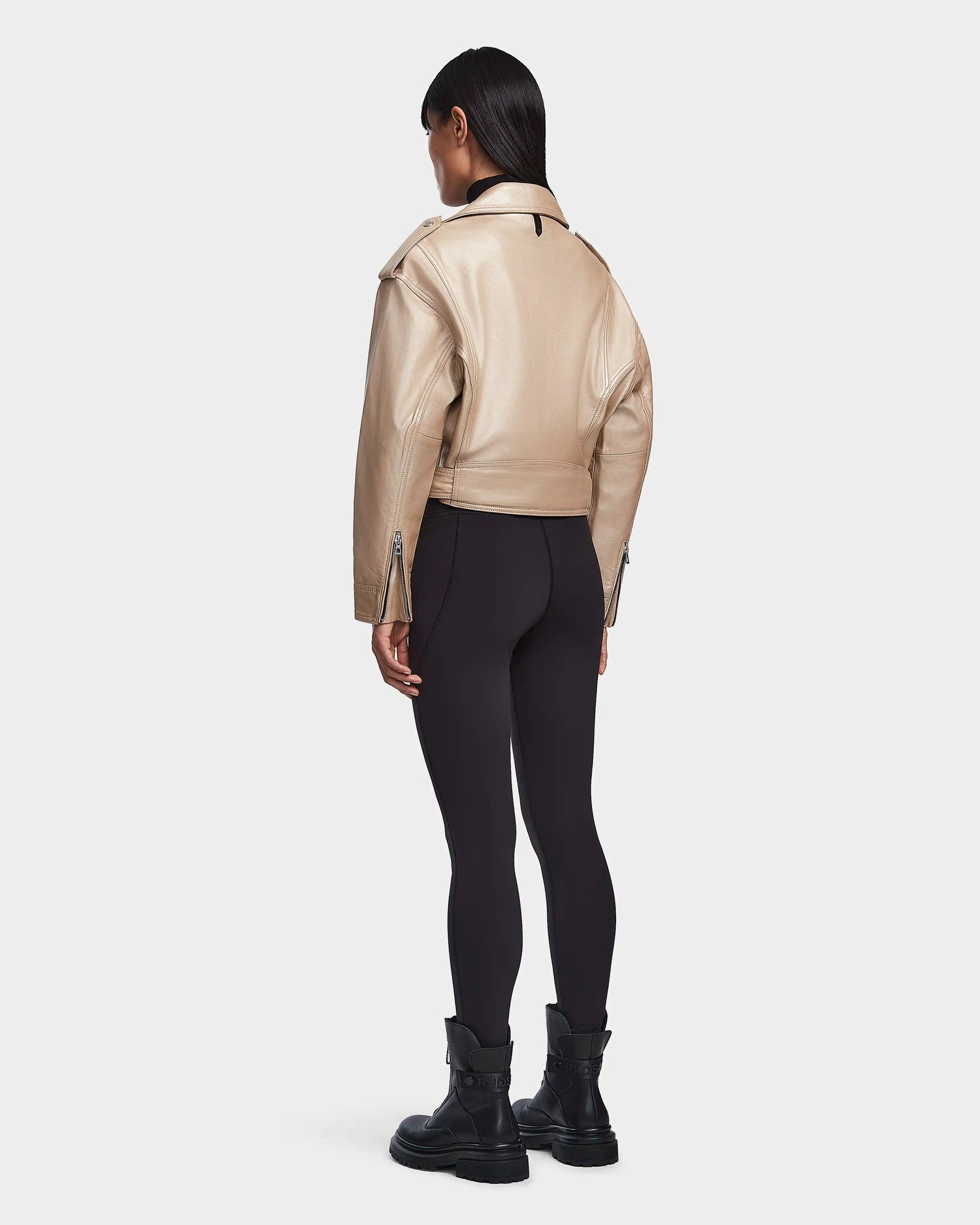 A person stands facing away, wearing the Rudsak MAGGIE - 8123031 Metallic Ivory cropped leather biker jacket, with black leggings and boots, against a plain light background.