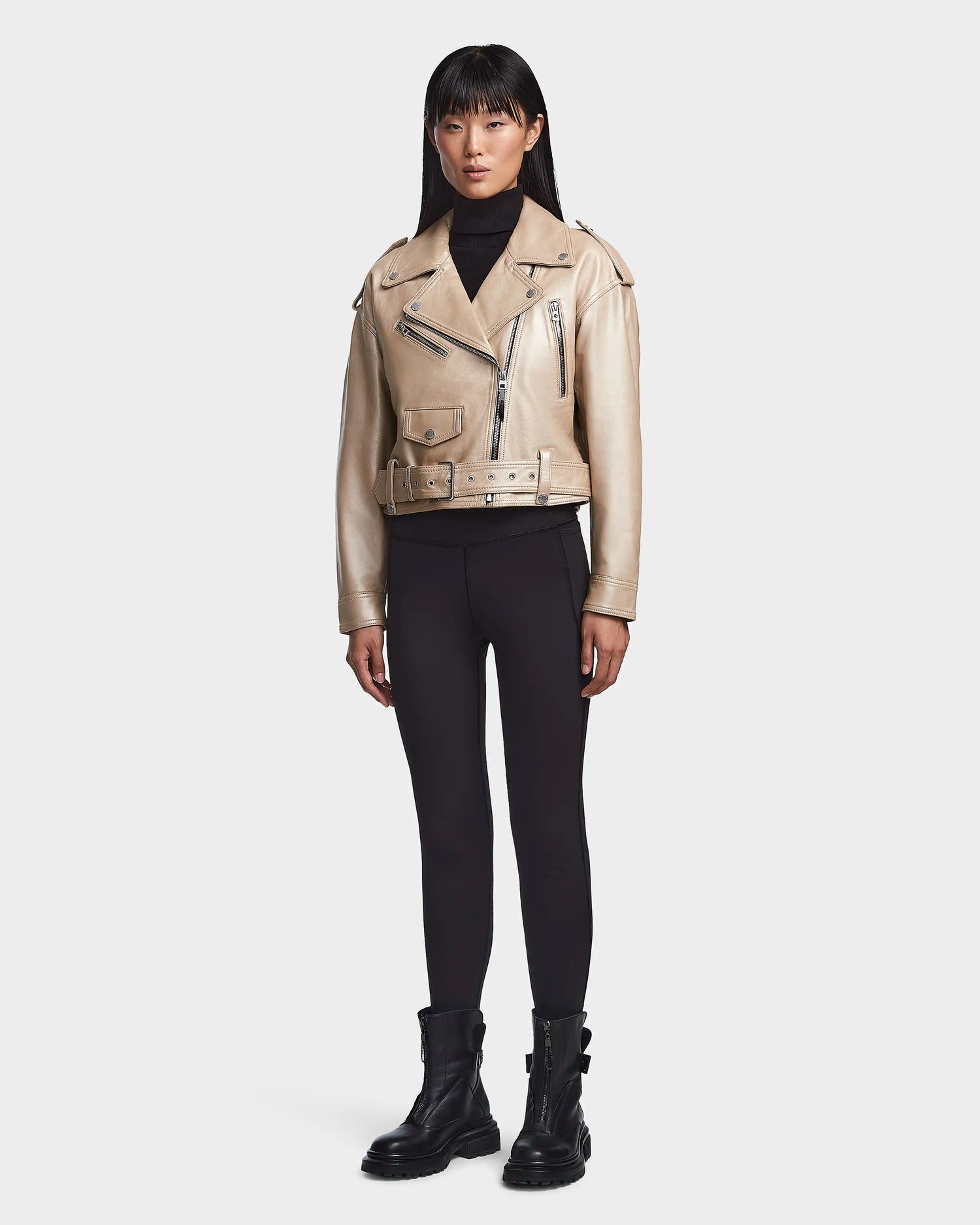 A woman faces forward against a white background, wearing the Rudsak MAGGIE - 8123031 Metallic Ivory cropped leather biker jacket with multiple zippers, black fitted pants, a black turtleneck, and chunky black ankle boots.