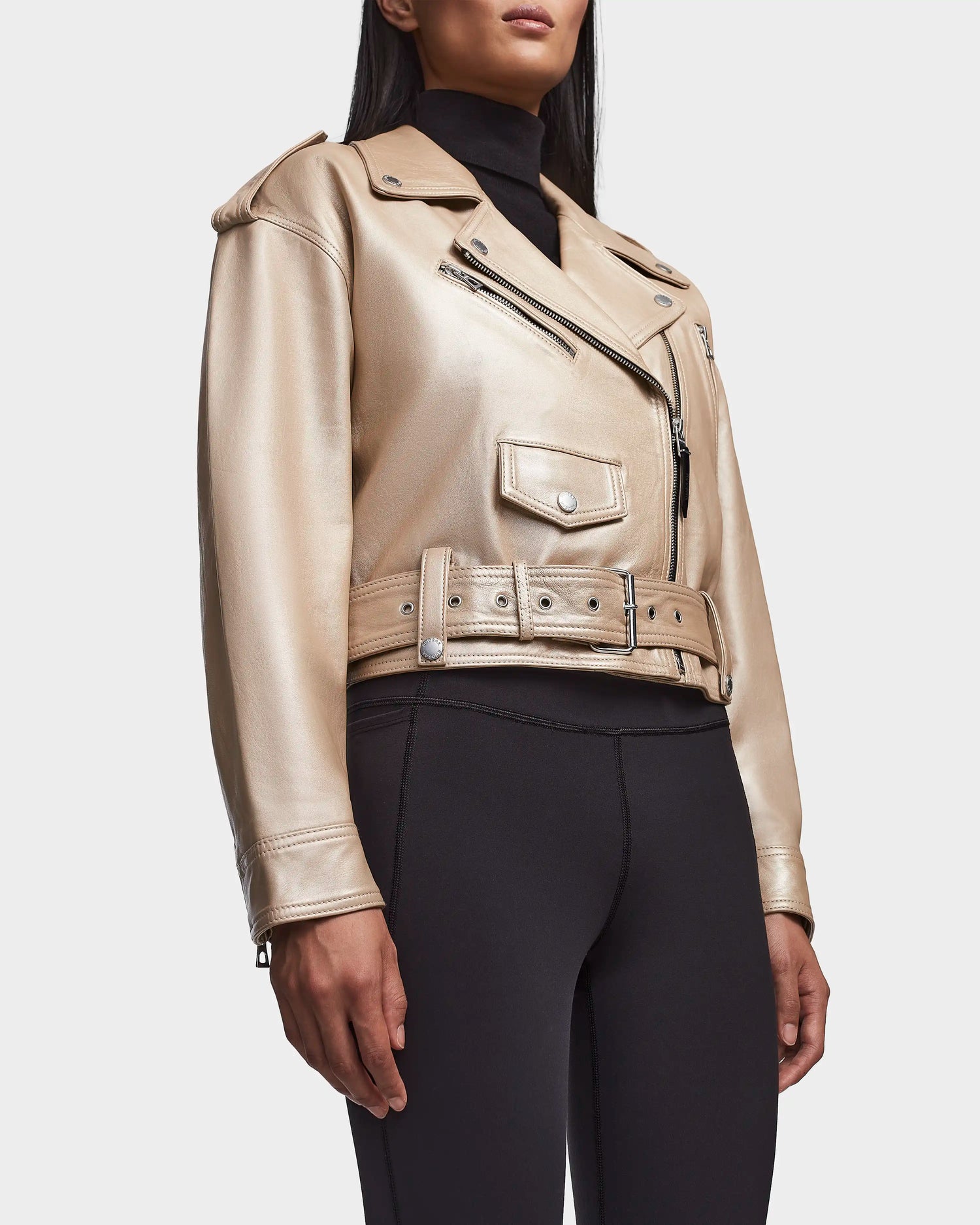 A person wears the Rudsak MAGGIE - 8123031 Metallic Ivory, a shiny gold cropped leather biker jacket with a belted waist and zippered pockets, paired with black fitted pants, standing against a plain white background.