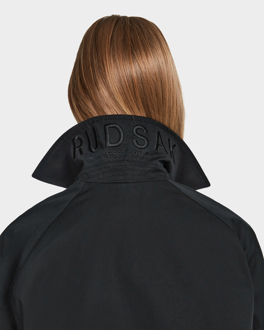 A person with straight, light brown hair is seen from behind wearing the Rudsak MIRELLE - 8124055 Black jacket. The tailored fit and embossed RUDSAK EST. 1994 on the upturned collar highlight its versatile style.