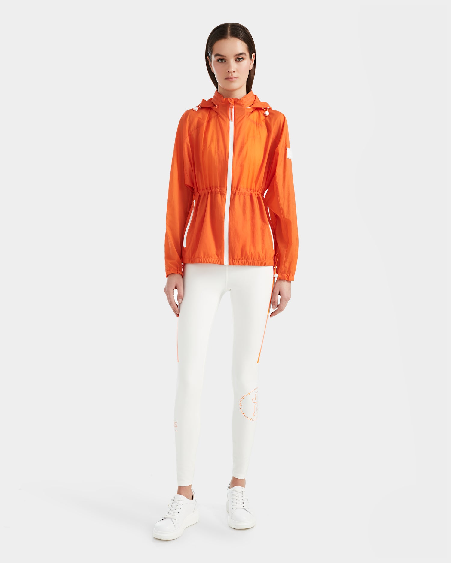 A woman faces forward against a white background, wearing the Rudsak WENDY - 8124009 Orange Sorbet lightweight jacket with a front zipper, paired with white fitted pants and sneakers.