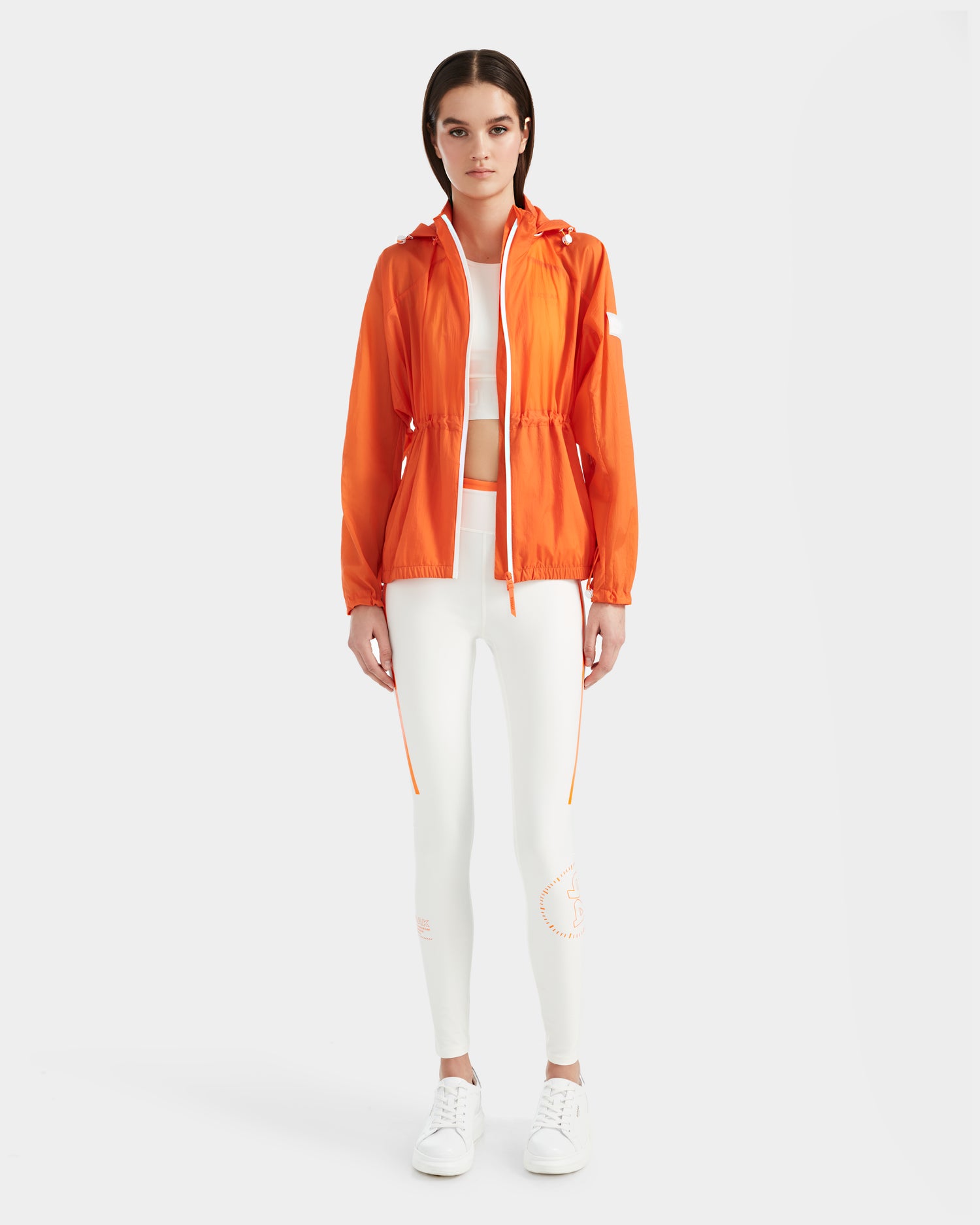 A woman models the Rudsak WENDY - 8124009 Orange Sorbet windbreaker over a white crop top and high-waisted leggings with orange accents, paired with white sneakers, standing against a plain white background.
