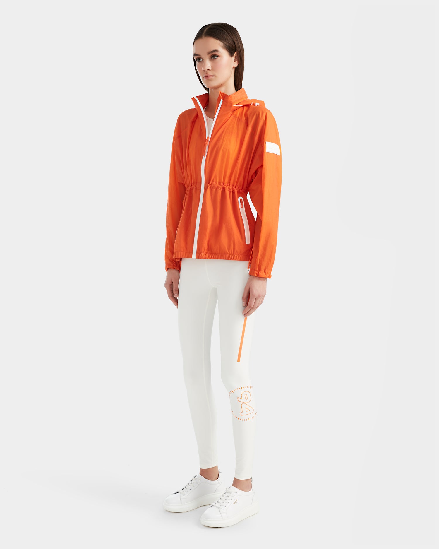 A woman poses against a white background in the Rudsak WENDY - 8124009 Orange Sorbet windbreaker with white accents, paired with white leggings featuring an orange and white patch, and white sneakers.