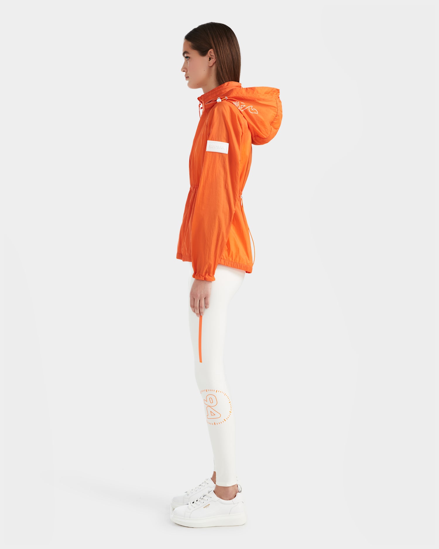A woman stands in profile wearing the Rudsak WENDY - 8124009 Orange Sorbet windbreaker, white leggings with orange graphics, and white sneakers against a plain white background.