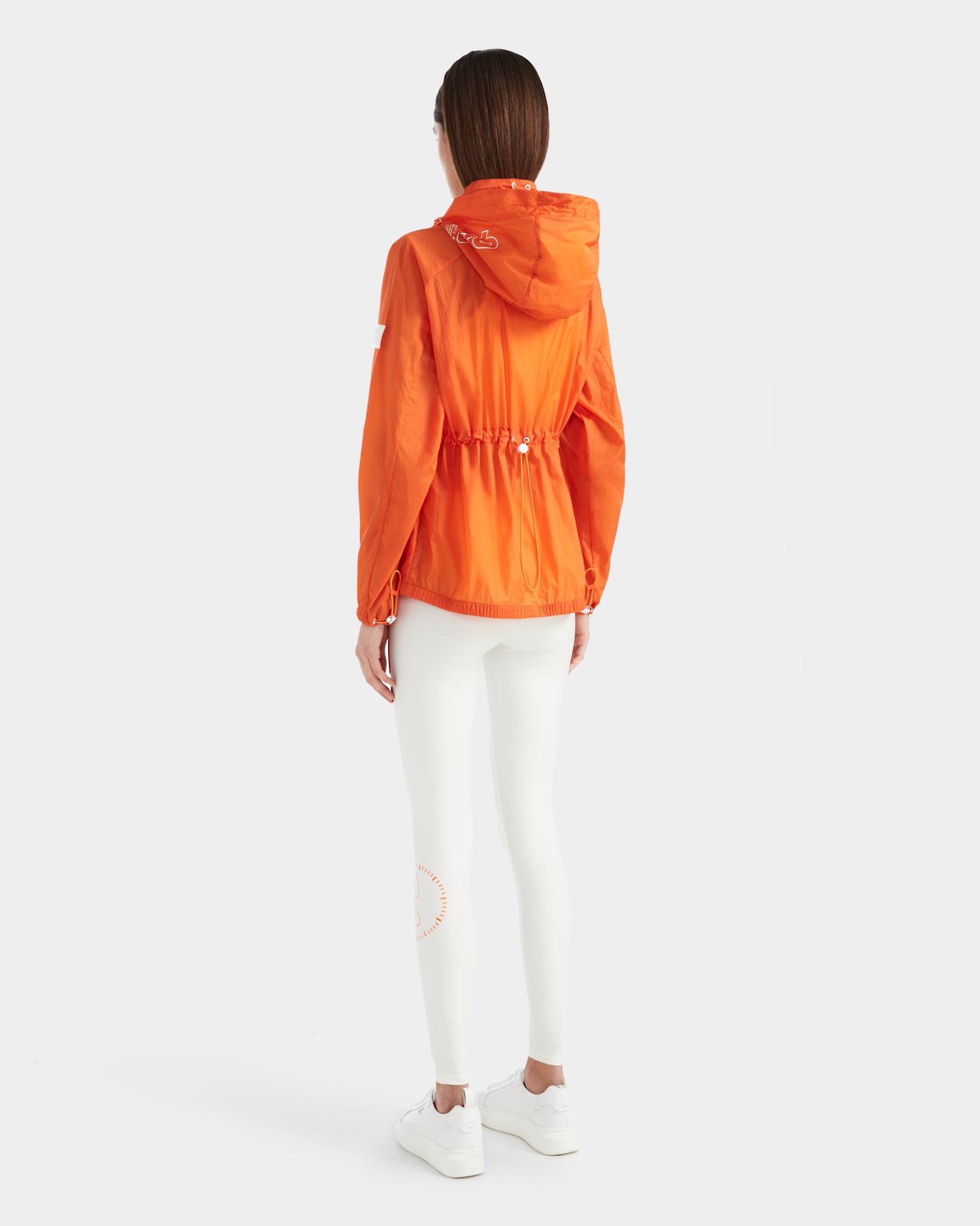 A person with straight brown hair is seen from behind, wearing the Rudsak WENDY - 8124009 Orange Sorbet women's windbreaker, paired with white leggings and sneakers, standing against a plain white background.
