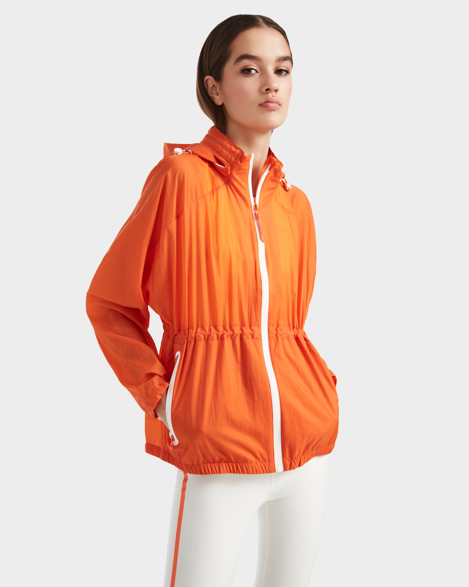 A woman models the Rudsak WENDY - 8124009 Orange Sorbet windbreaker with white zipper and drawstrings, styled with white pants against a light gray background. This lightweight jacket is ideal for both activewear and casual looks.