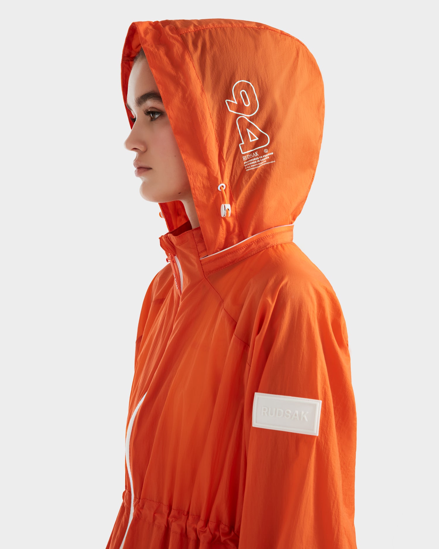 A person in profile wears the Rudsak WENDY - 8124009 Orange Sorbet women's windbreaker, featuring white drawstrings and logo details on the hood and sleeve, standing against a plain light background.