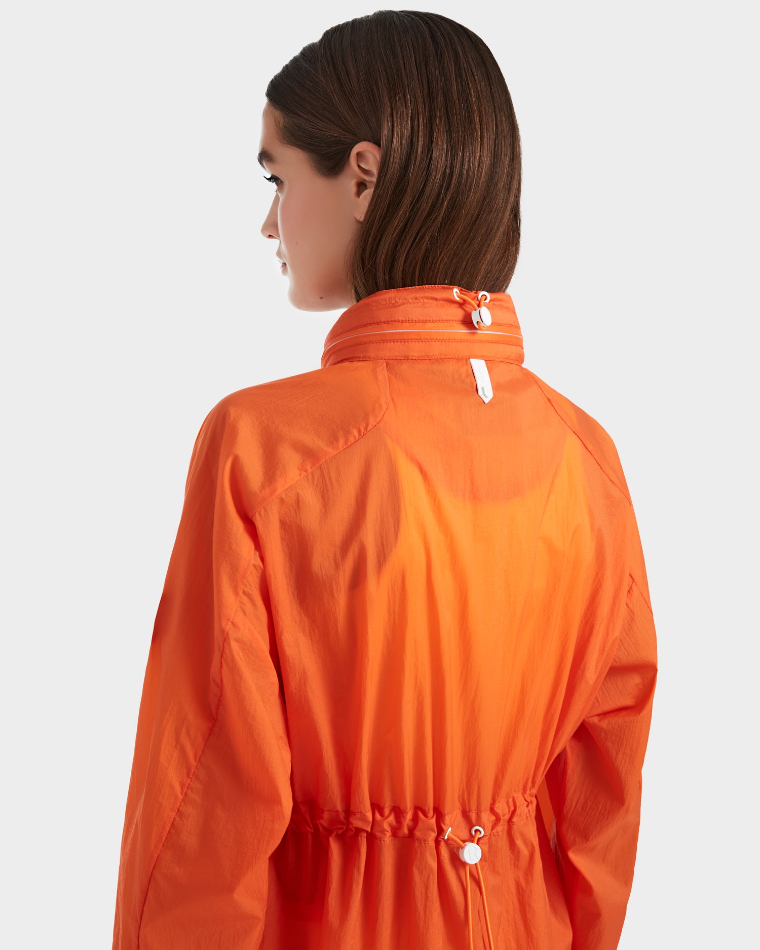 Seen from the back, a person with straight brown hair wears the Rudsak WENDY - 8124009 Orange Sorbet women’s windbreaker, featuring a high collar and adjustable drawstrings, standing out against a plain light gray background.