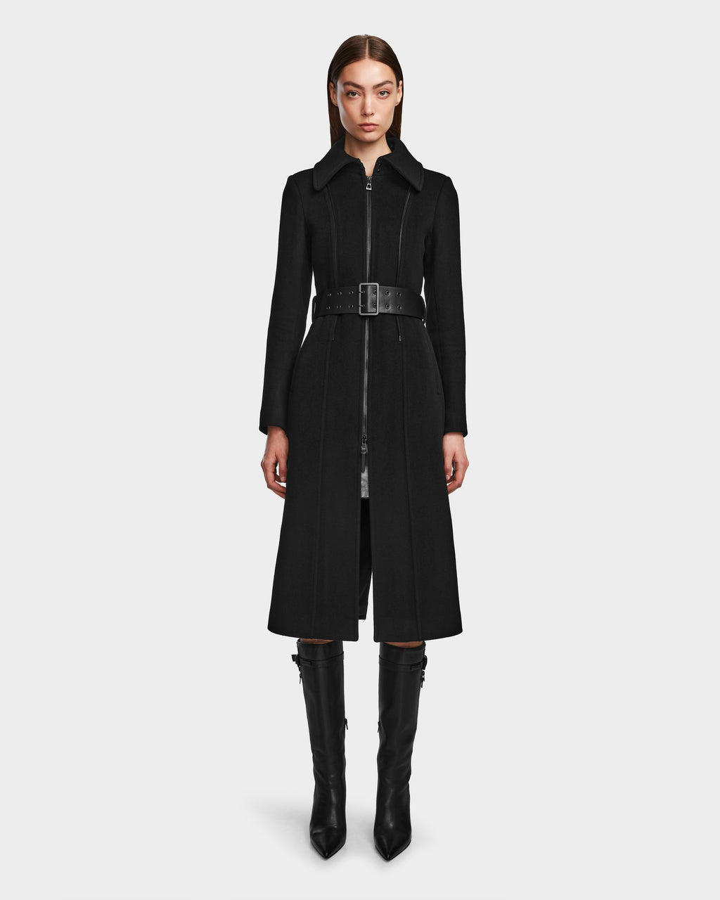 A woman stands facing forward, wearing the Rudsak VIRGINIE - 8124696 Black women’s long peacoat with a zippered front and pointed collar, styled with knee-high black boots. The coat features a removable faux fur collar for added flair against a plain white background.