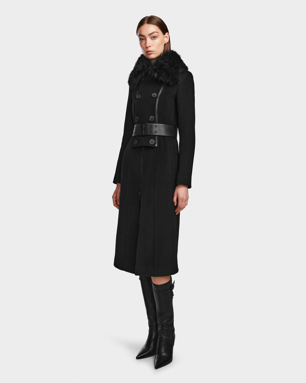A woman wears the Rudsak VIRGINIE - 8124696 Black peacoat, crafted from a wool-cashmere blend with a removable faux fur collar and belted waist, styled with black knee-high heeled boots against a plain background.