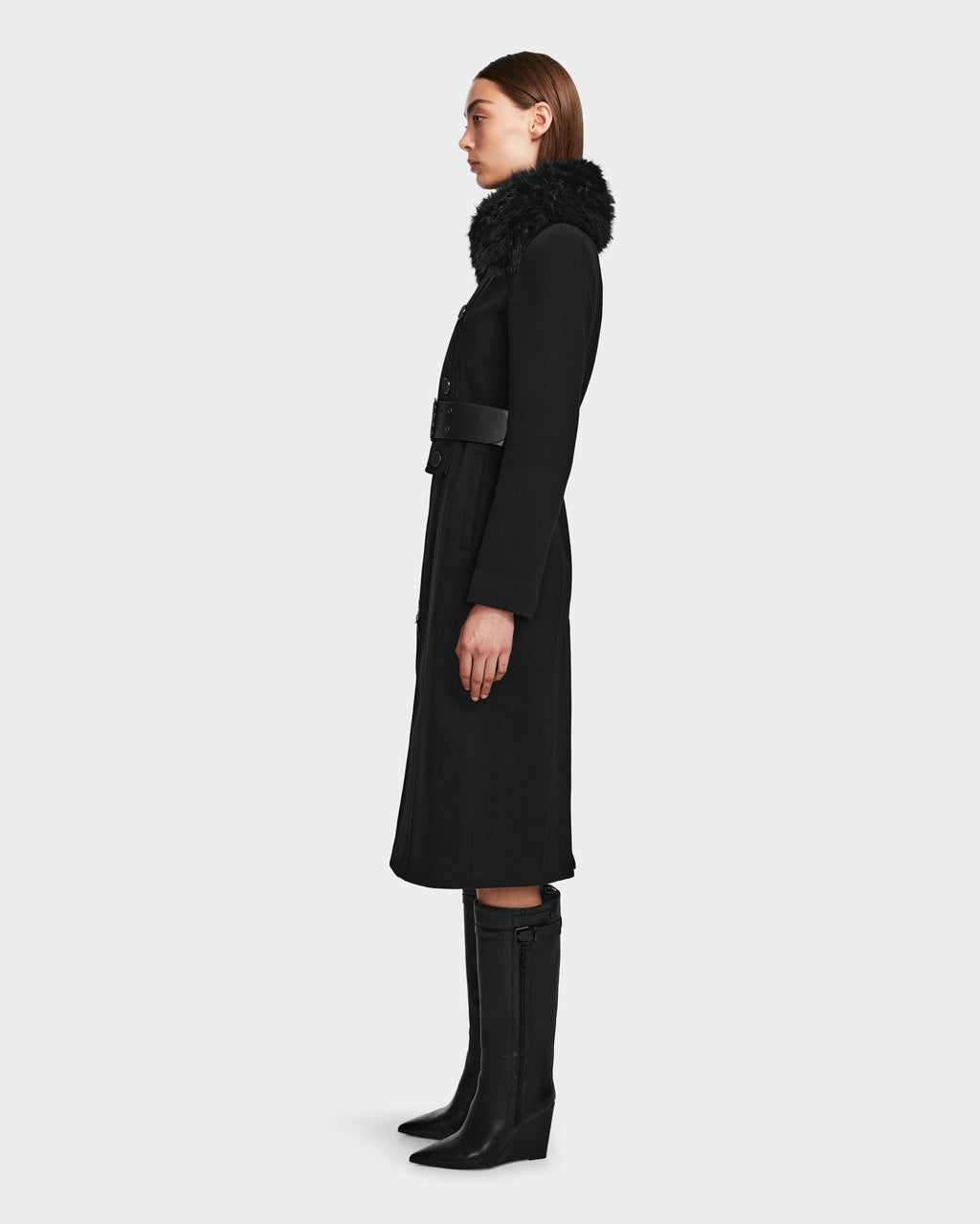 A woman stands in profile wearing the Rudsak VIRGINIE - 8124696 Black wool and cashmere coat with a removable faux fur collar, belted waist, and knee-high black boots against a plain light background.