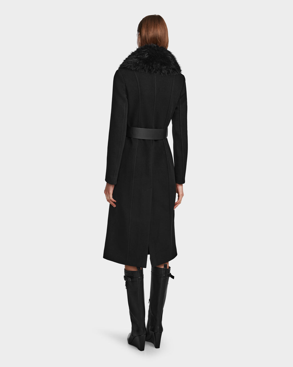 A person with straight, shoulder-length hair stands facing away, wearing the Rudsak VIRGINIE - 8124696 Black women’s long peacoat with a wide belt and removable faux fur collar, paired with tall black boots.