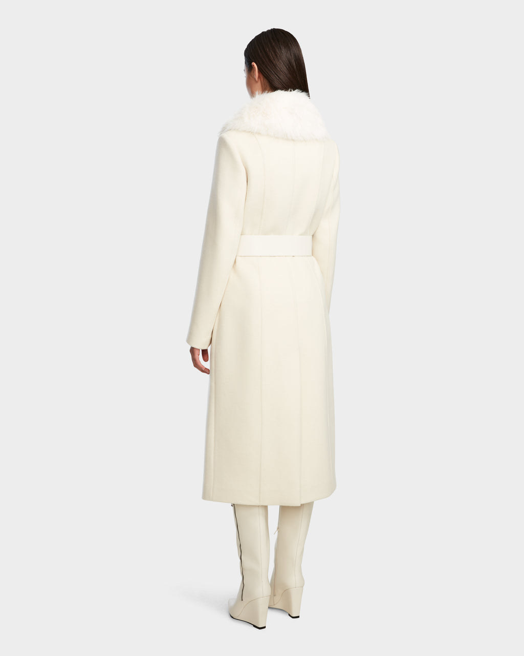 A woman stands facing away, wearing the Rudsak VIRGINIE - 8124696 Iceland long peacoat with a wide white belt and removable faux fur collar, paired with matching white pants and pointed-toe boots against a plain light gray background.