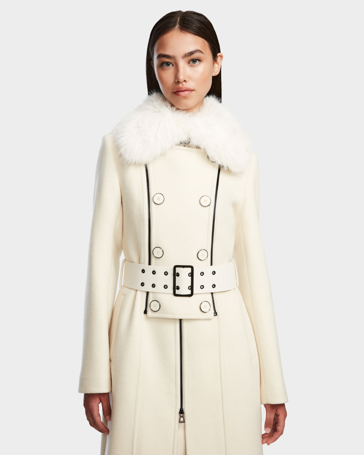A woman models the Rudsak VIRGINIE - 8124696 Iceland womens long cream peacoat with a removable faux fur collar, black trim, double-breasted buttons, and a belted waist against a light gray background.