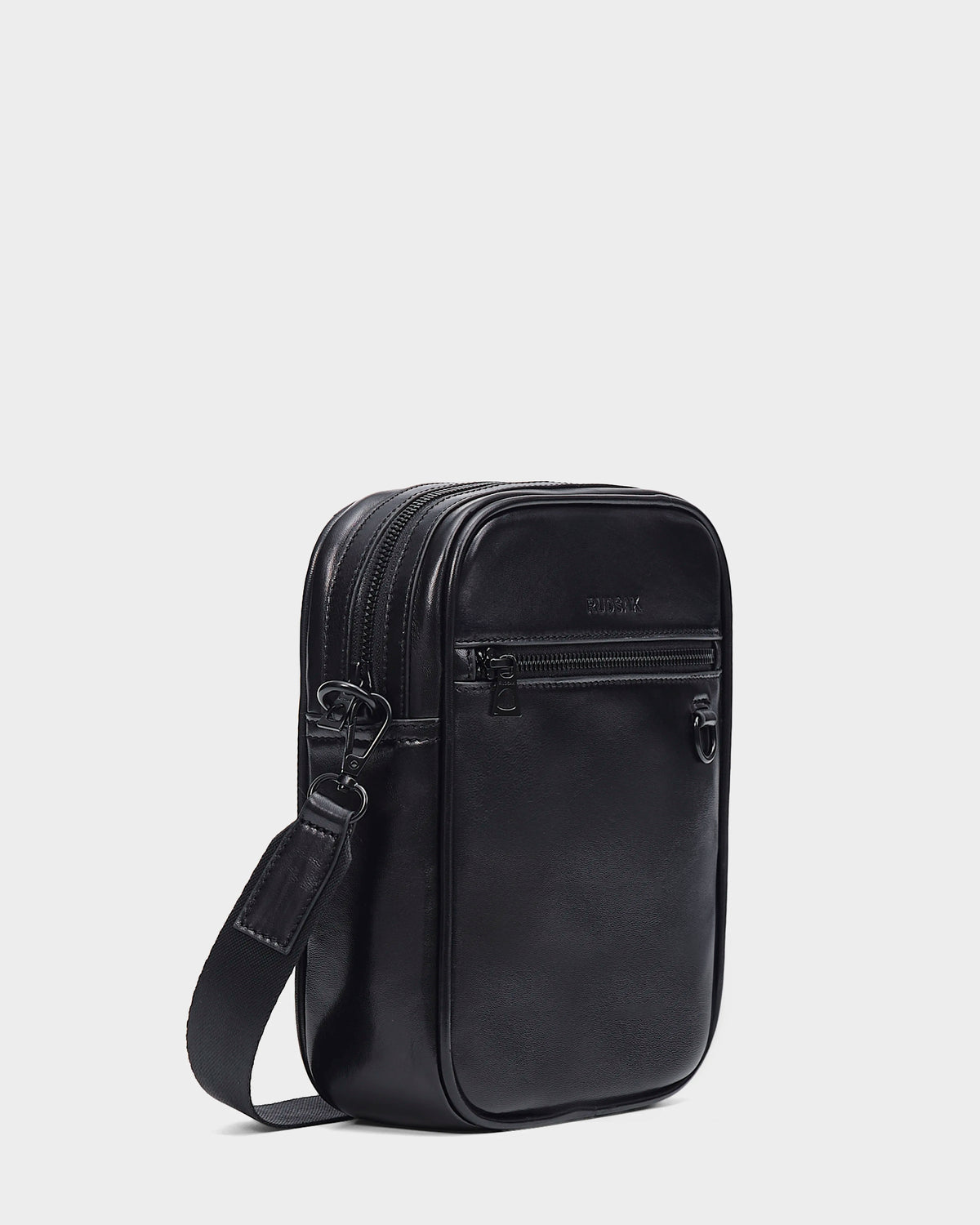 The Rudsak NORMAN L - 9323503 Black is a small, rectangular unisex crossbody bag in black leather with two zippered compartments, a front zip pocket, and an adjustable strap, shown against a plain white background.
