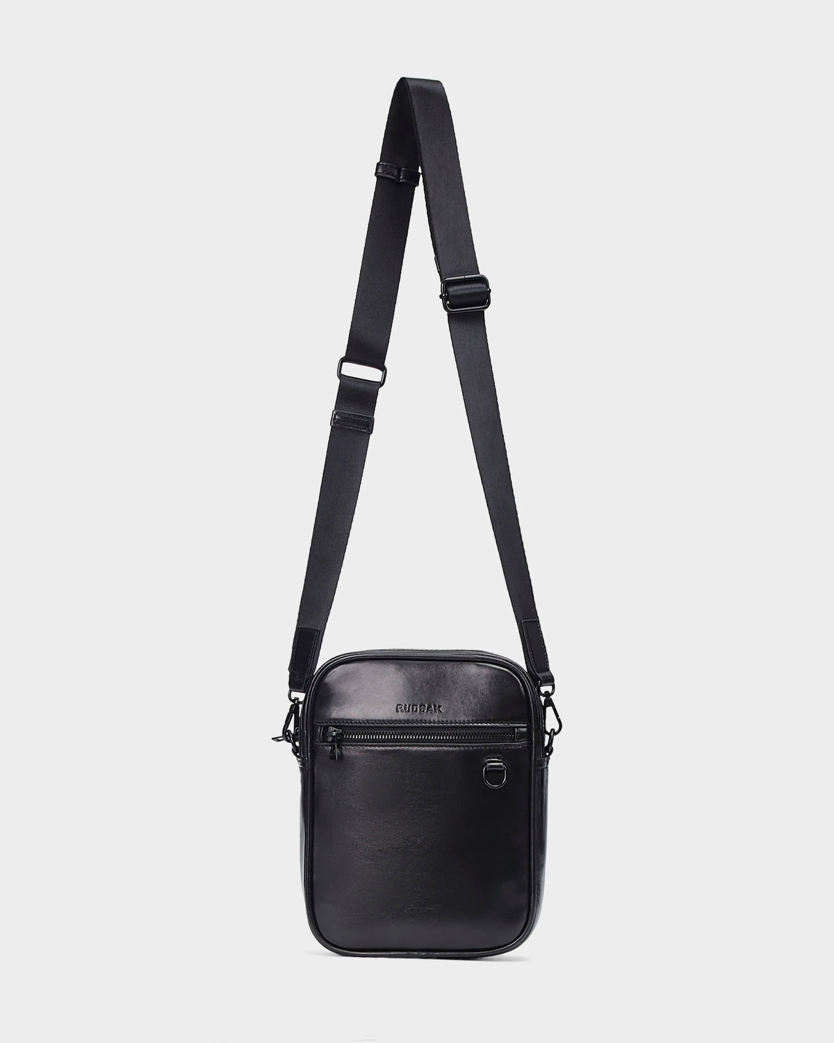 The Rudsak NORMAN L - 9323503 Black is a unisex black leather crossbody bag with an adjustable strap, front zipper pocket, and metal ring detail, shown against a plain white background.