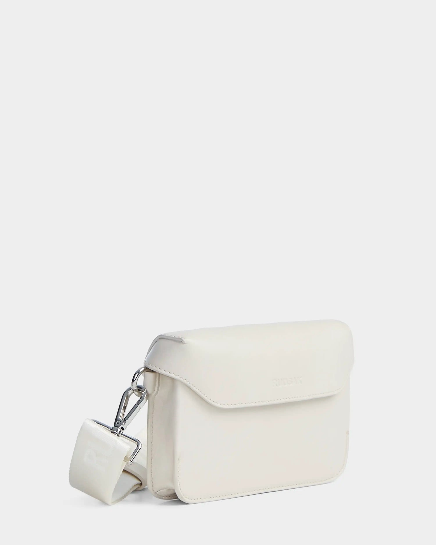 The CAMELIA - 8324000 Eggshell by Rudsak is a women’s leather crossbody bag with a rectangular white design, flap closure, silver clasp, and matching wide strap, shown on a plain white background.
