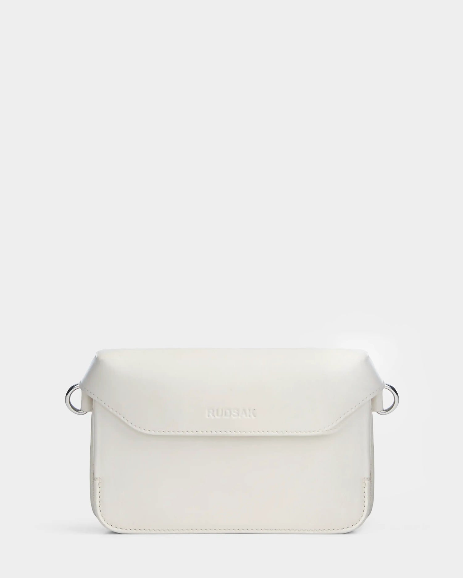 The CAMELIA - 8324000 Eggshell by Rudsak is a minimalist women’s leather crossbody in white, featuring a flap closure, silver strap rings, and an embossed logo on the front, shown against a plain light gray background.