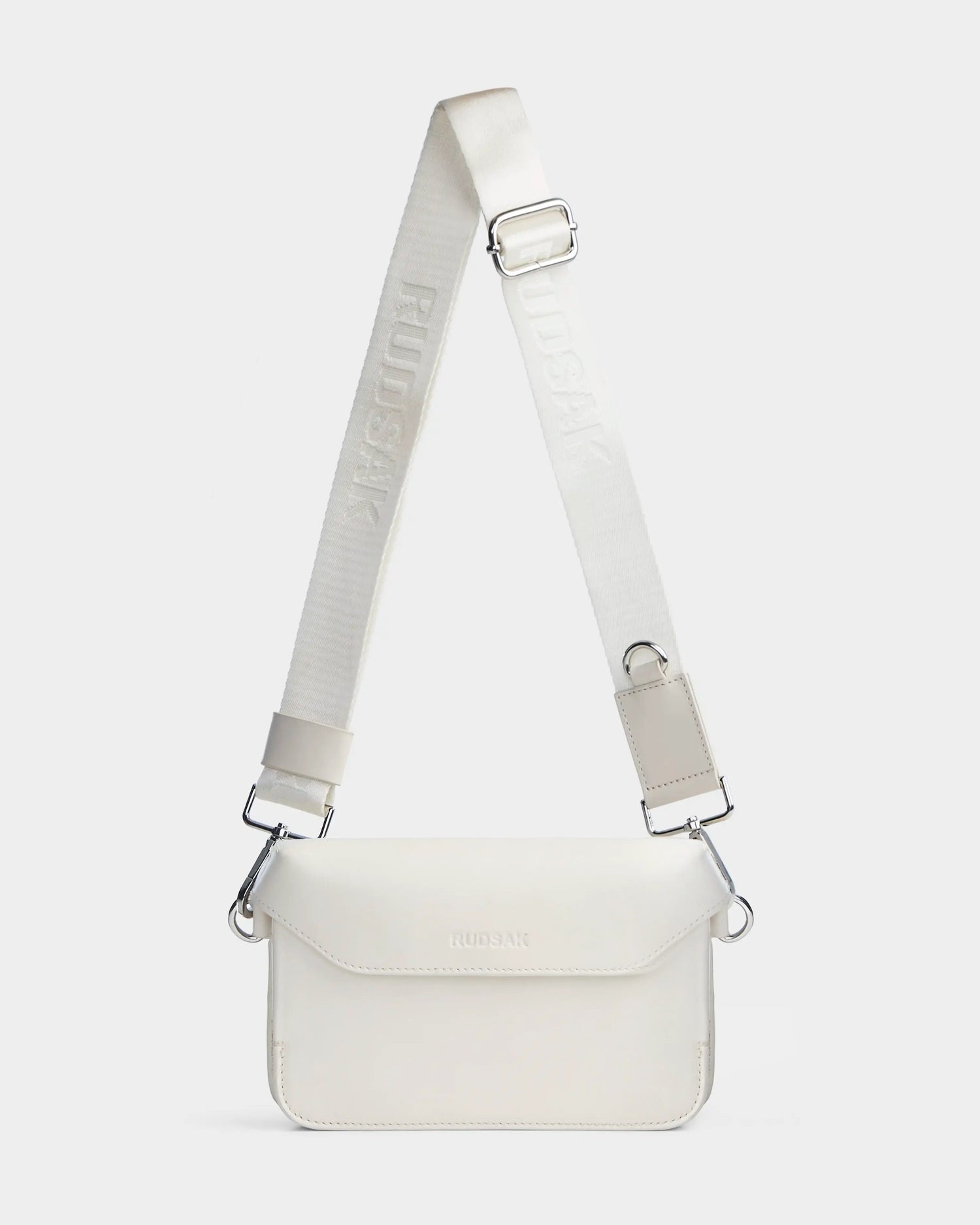 CAMELIA - 8324000 Eggshell by Rudsak is a minimalist women’s leather crossbody bag with a rectangular shape, flap closure, silver hardware, and a wide adjustable branded strap. Shown against a plain light gray background.