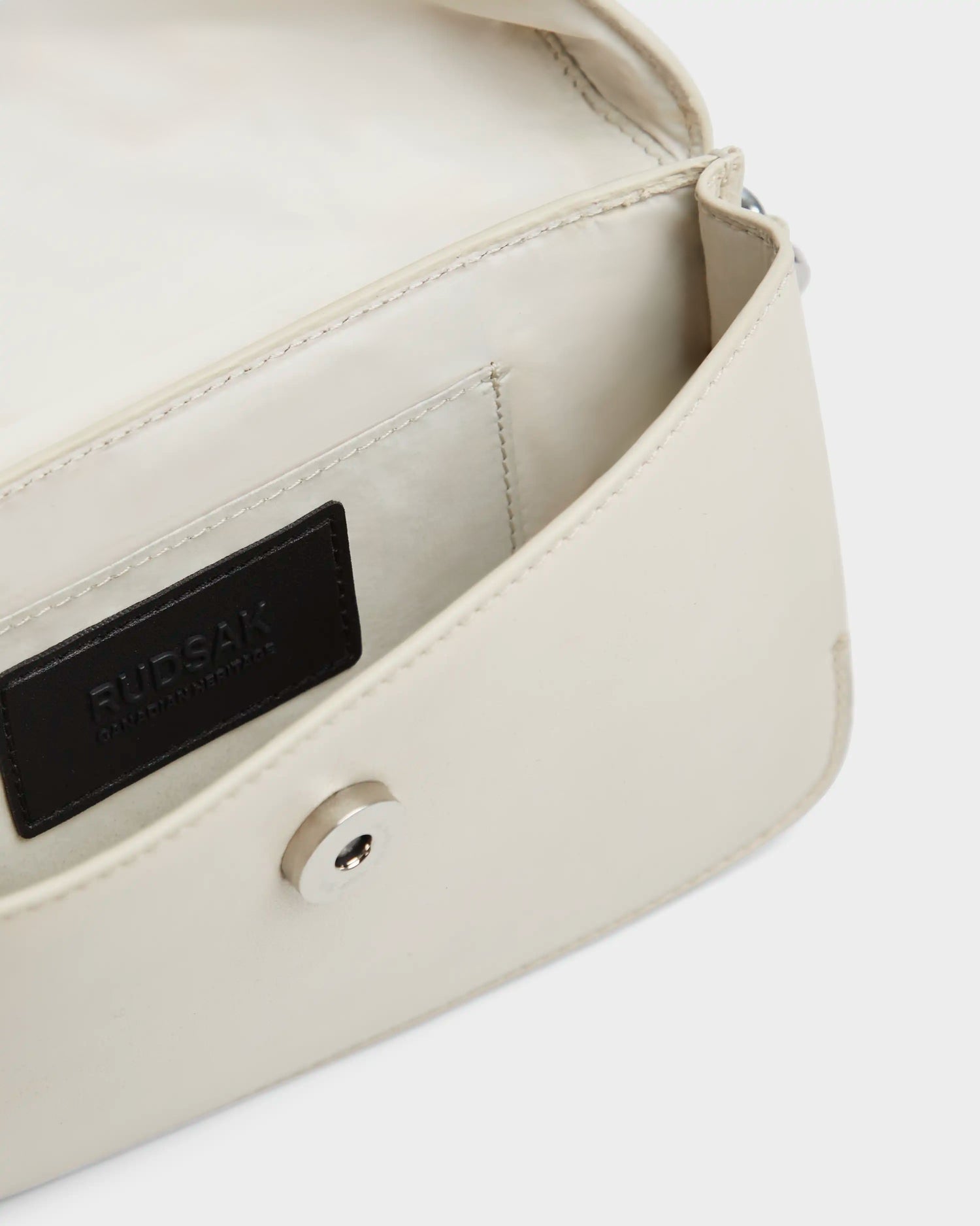 The CAMELIA - 8324000 Eggshell by Rudsak is a women’s leather crossbody bag featuring a flap opening, a light interior pocket, and a black brand label. This small bag is completed with a silver snap button closure.