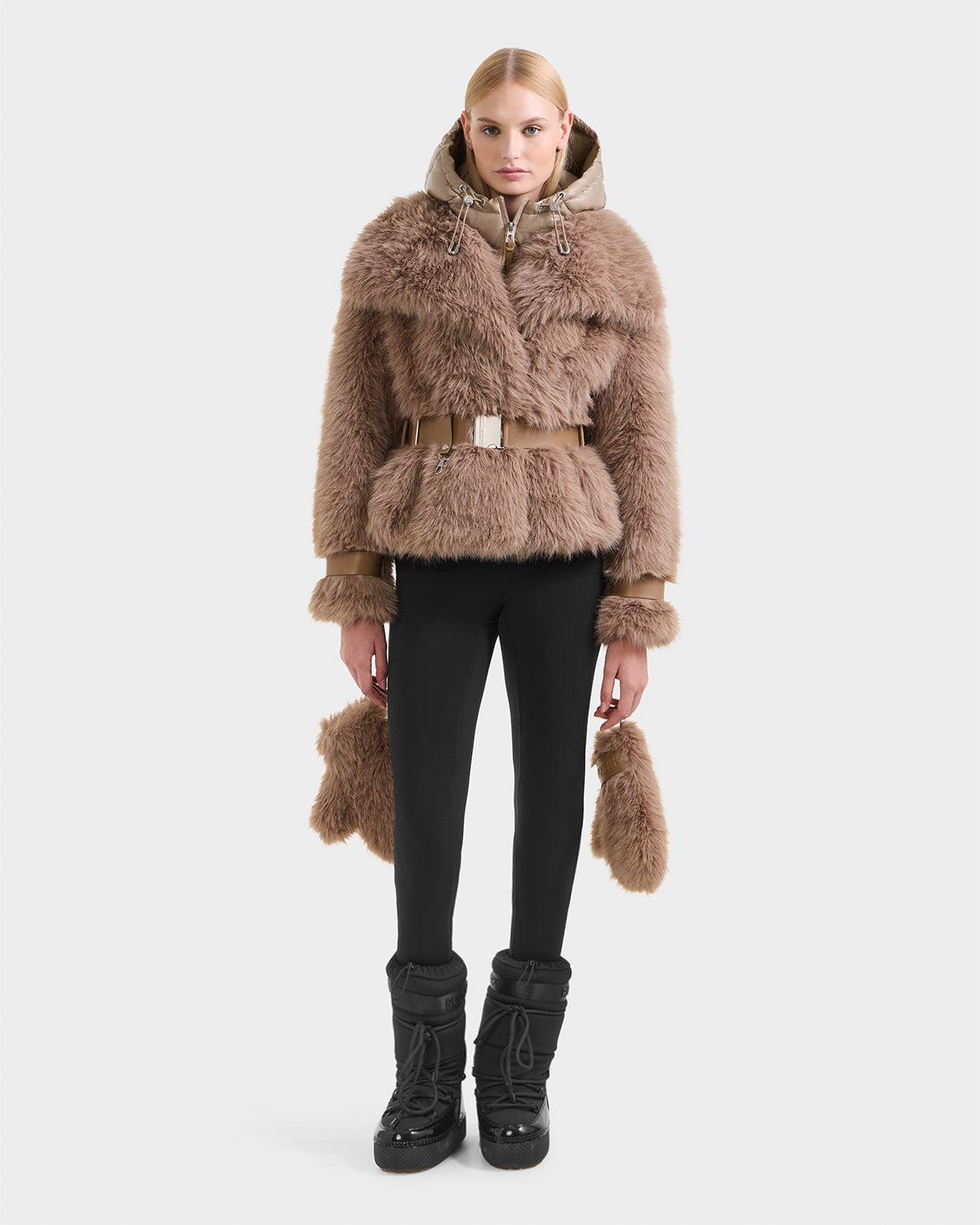 A woman with straight blonde hair and a neutral expression faces forward, wearing the CHLOE - 8125619 Camel women's faux fur jacket by Rudsak, paired with black leggings and winter boots. The background is plain white.