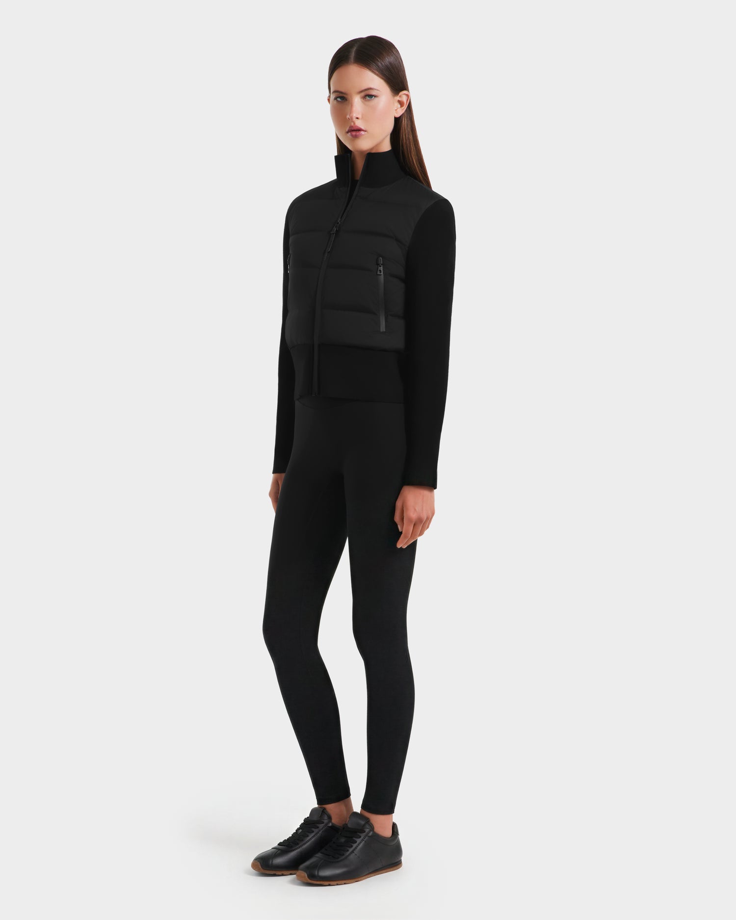 A woman stands against a plain white background, wearing the Rudsak CLAREMONT - 8125502 Black women's hybrid down jacket for lightweight warmth, paired with black leggings and shoes. She faces forward with her arms relaxed at her sides.