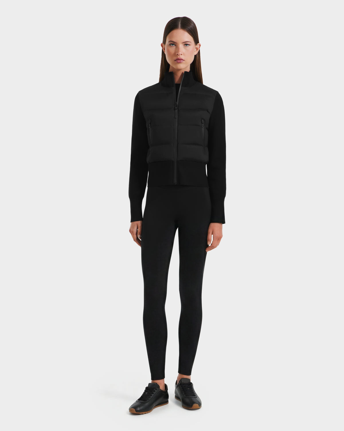 A woman faces forward against a plain white background, wearing Rudsak's CLAREMONT - 8125502 Black zip-up padded vest, paired with a black long-sleeve top, black leggings, and black sneakers.