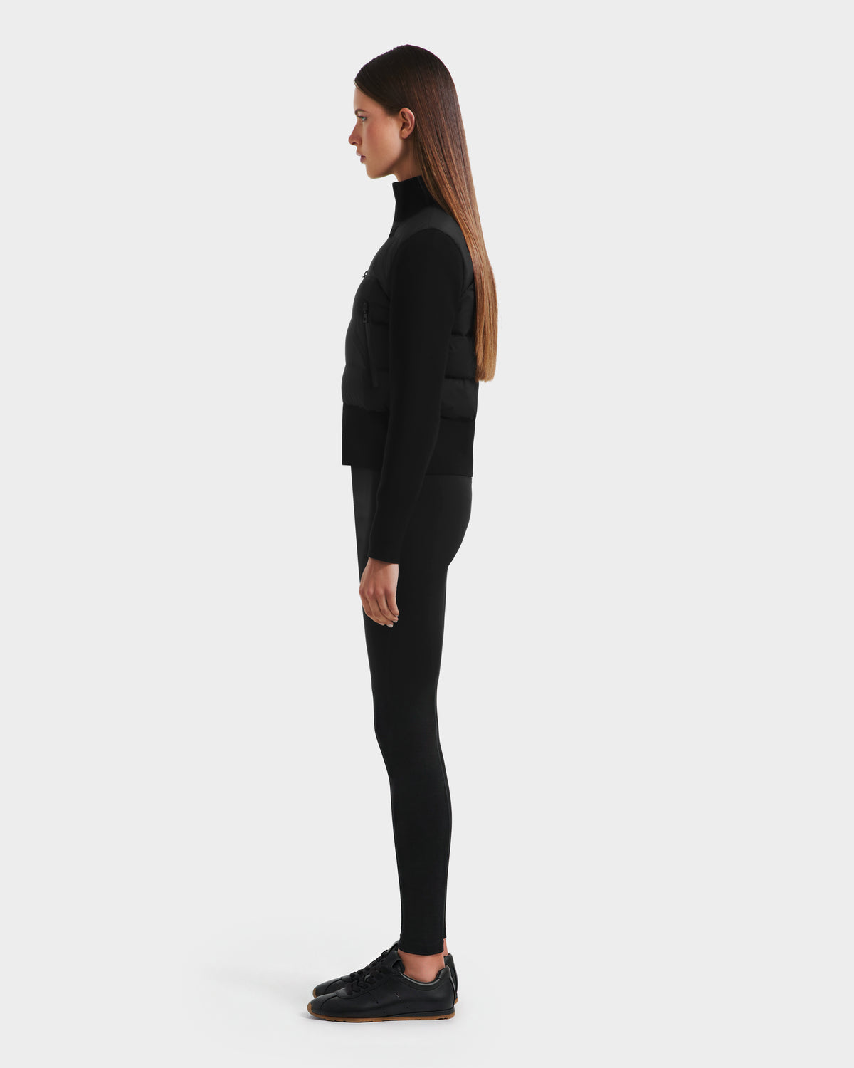 A woman stands in profile against a white background, wearing the Rudsak CLAREMONT - 8125502 Black women's hybrid down jacket, which offers lightweight warmth with RDS-certified down. Her look includes a black turtleneck, vest, and leggings.