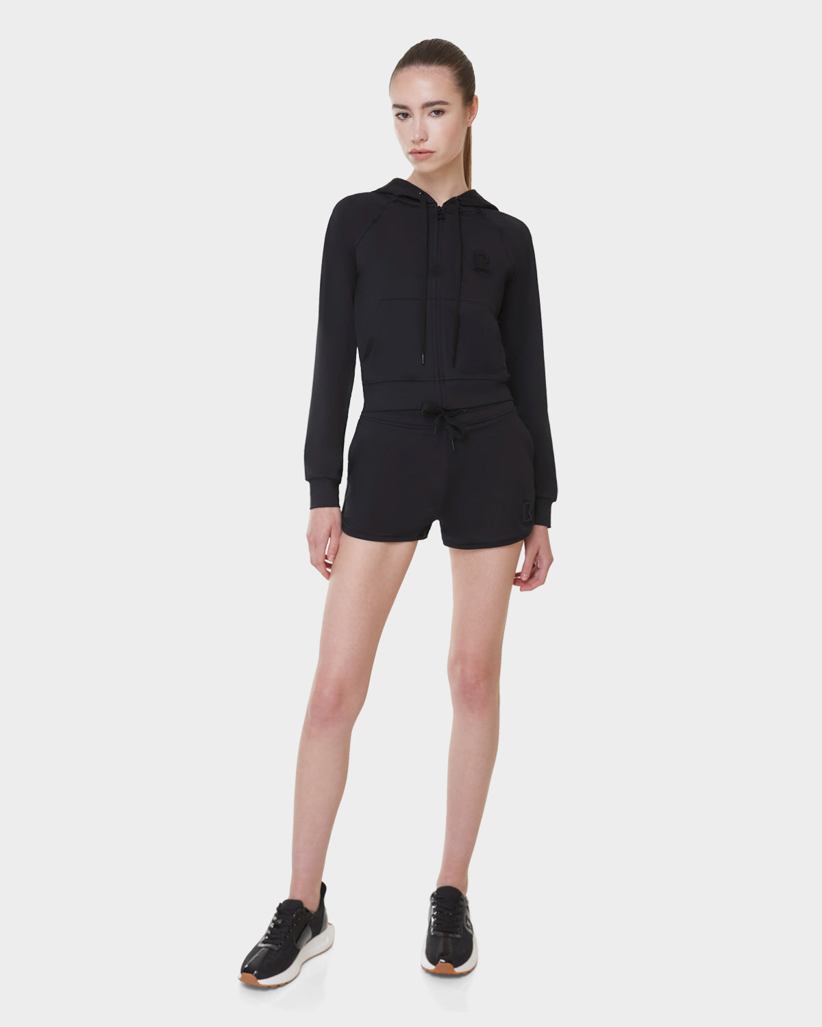 A woman stands against a plain light background wearing Rudsak's SAMMY - L523014B bottoms in black, paired with a black zip-up hoodie and black sneakers with white soles. Her straight hair is tied back and her arms are relaxed by her sides.