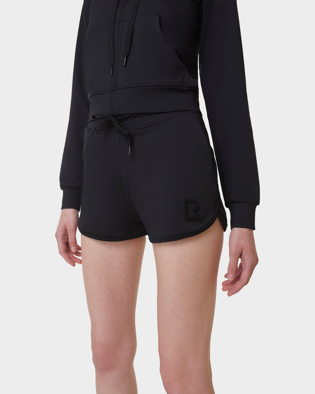 A person is wearing Rudsak's SAMMY - L523014B black bottoms, featuring a small logo on the left leg, paired with a black zip-up hoodie, standing against a plain white background.