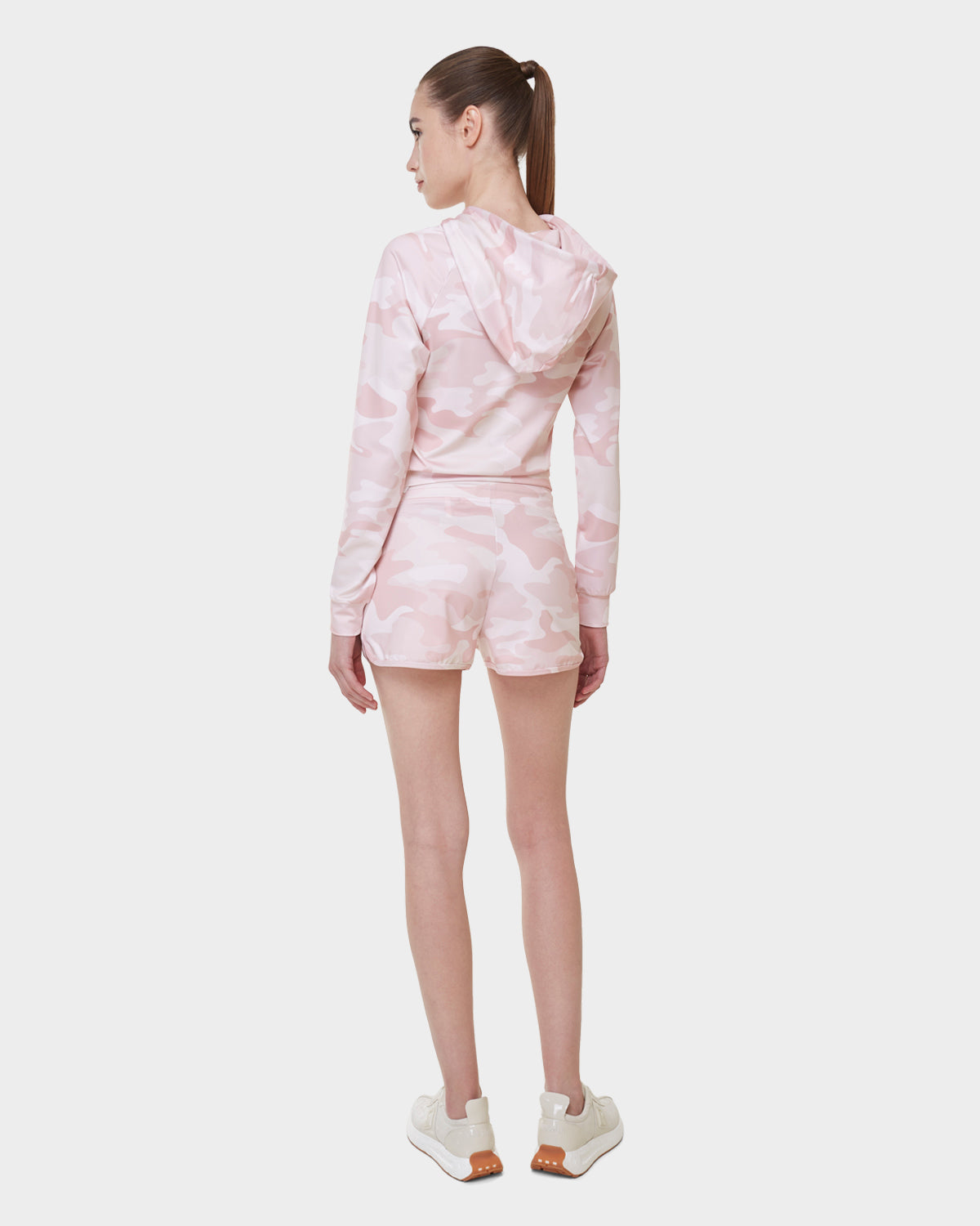 A woman with a ponytail faces away, wearing Rudsak's SAMMY - L523014B Camouflage Pink shorts and a matching hoodie with white sneakers against a plain white background.
