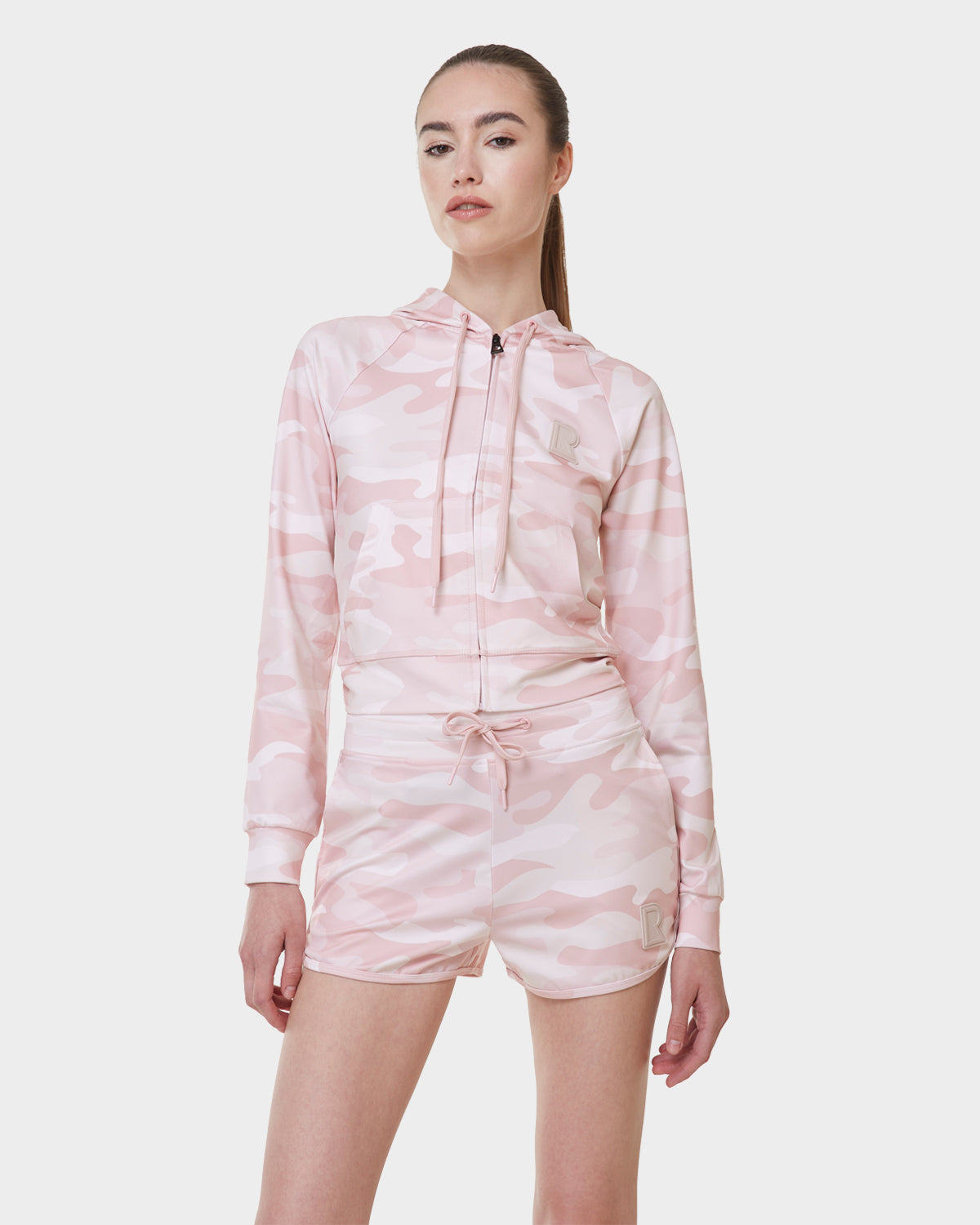 A woman with straight brown hair pulled back wears Rudsak’s SAMMY - L523014B Camouflage Pink drawstring shorts, paired with a matching hooded zip-up jacket, standing against a plain white background.