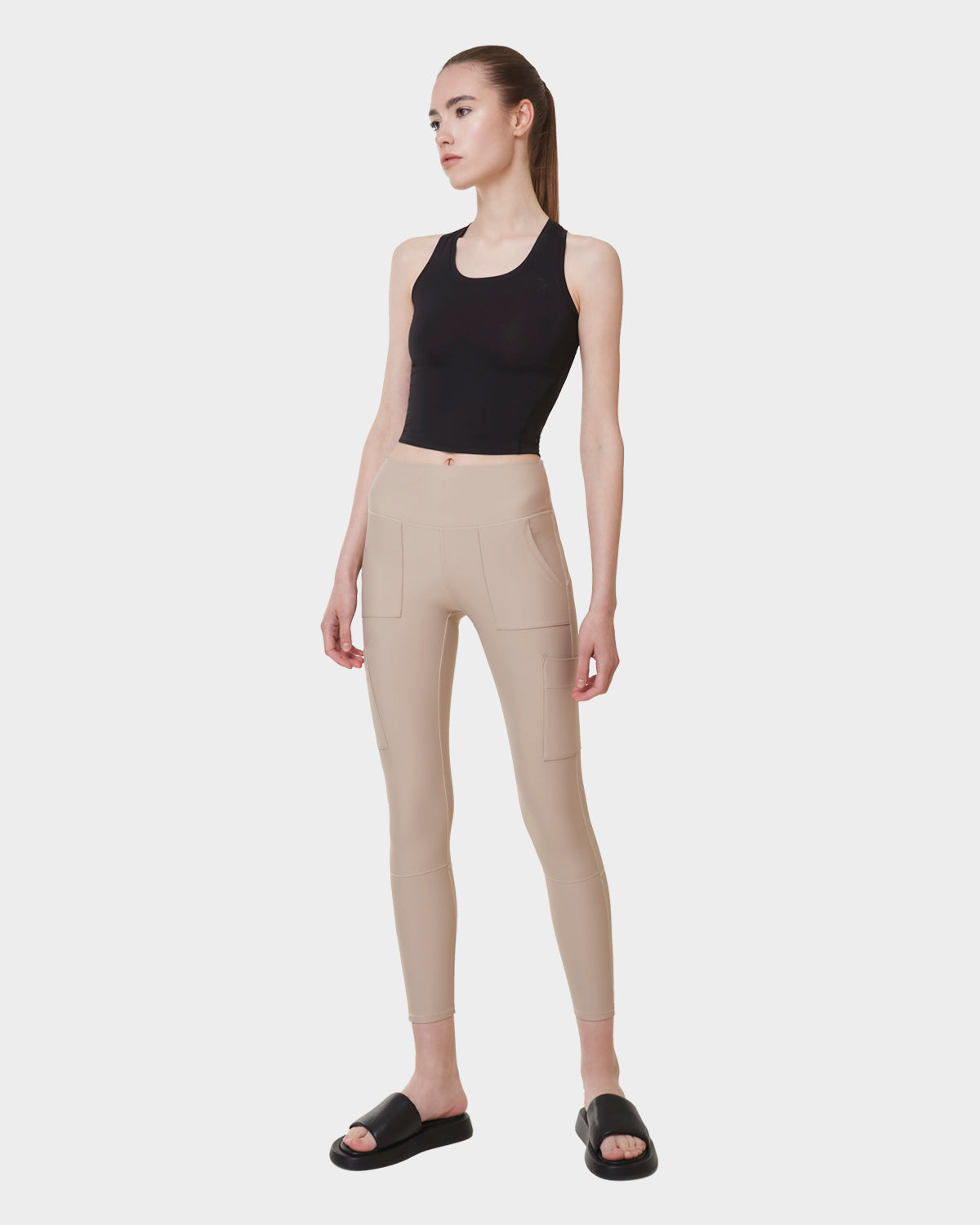 A woman stands against a white background wearing the Rudsak ADELLE - L523032B Cream high-waisted leggings with pockets, paired with a black crop top and black slide sandals. She gazes slightly to the side, arms relaxed.