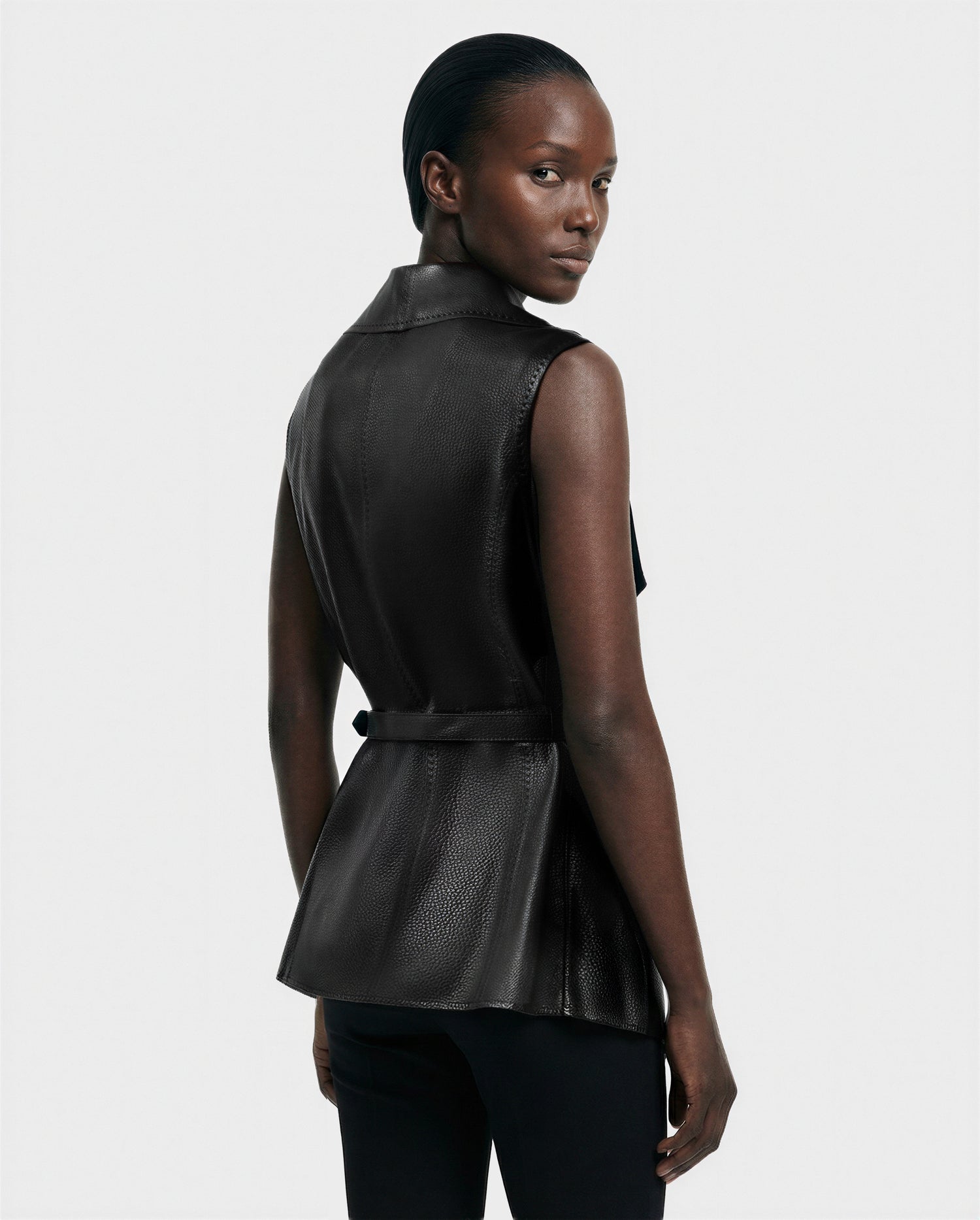 A person with dark, slicked-back hair models the Rudsak ELEANOR - 8125584 Black sleeveless textured leather vest with a belted waist, styled with black pants, posing against a white background and looking over their shoulder.