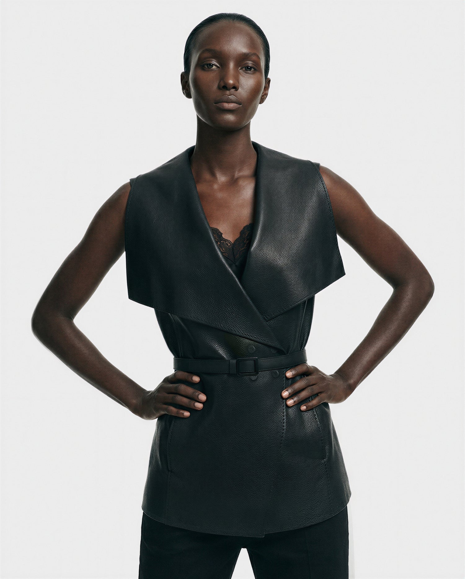A woman stands confidently with hands on hips, wearing the Rudsak ELEANOR - 8125584 Black women’s full-grain leather vest with a wide shawl collar and black belt over a lace-trimmed top, set against a plain white background.