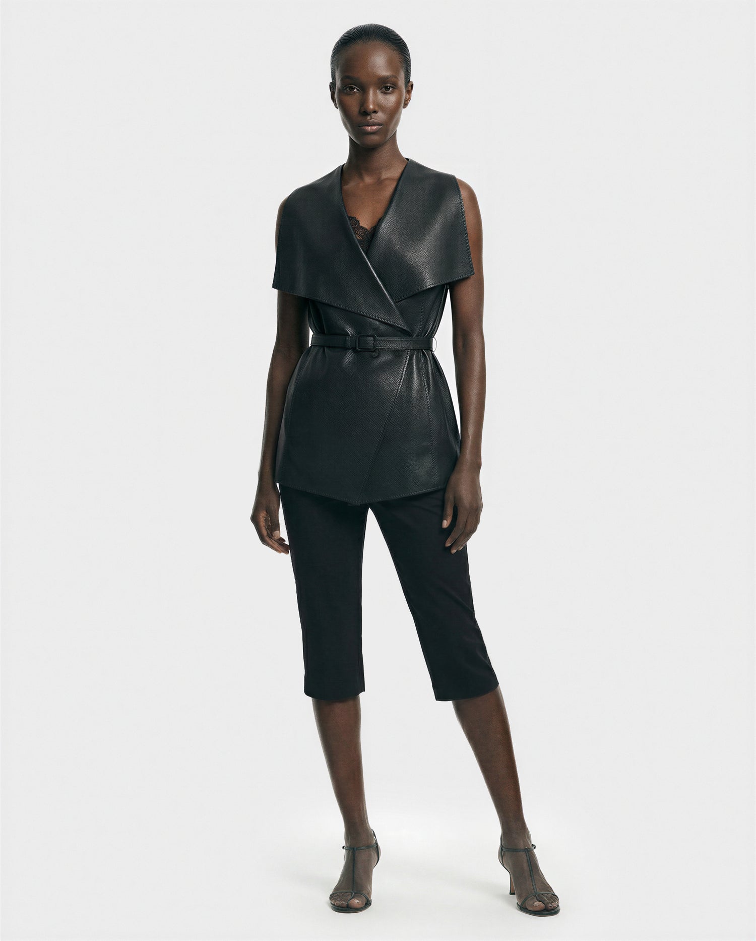 A woman poses against a plain light background wearing the Rudsak ELEANOR - 8125584 Black sleeveless leather vest with a belted waist, styled over a black top, cropped black pants, and open-toe heeled sandals.