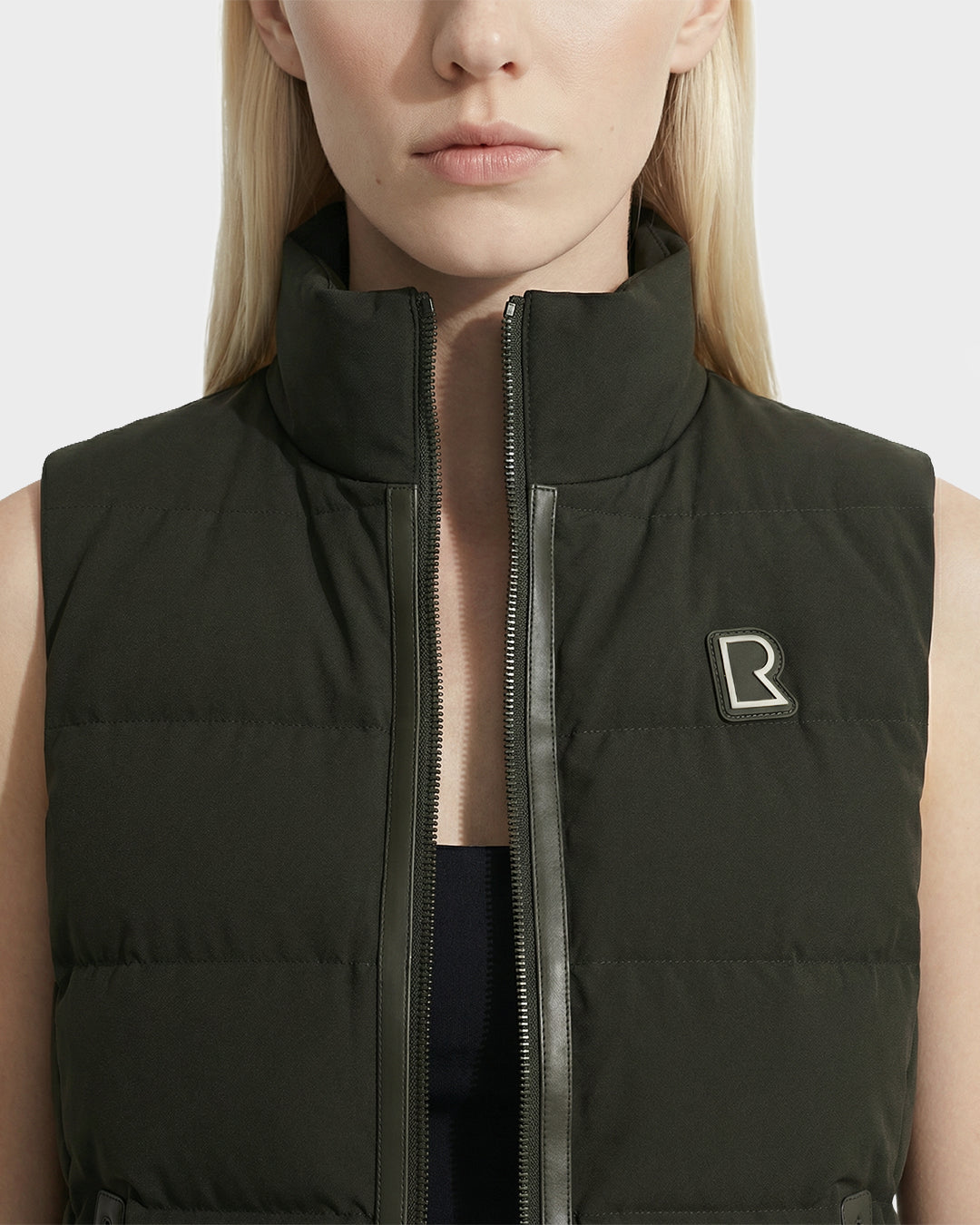 A woman wears the Rudsak GABRIELA - 8125014 Dark Olive vest with a stand collar, zippered front, and R logo patch on the chest. Her straight blonde hair frames her partially visible face.